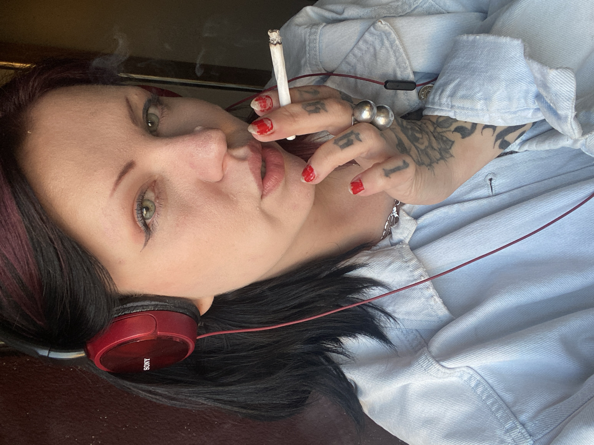 A woman is reclining and holding a lit cigarette near her lips. She has dark hair with red highlights, green eyes, and red nail polish. There are tattoos on her fingers and the back of her hand. She is wearing red Sony headphones and a light blue denim shirt. A silver necklace is visible. The location is not identifiable from the image.