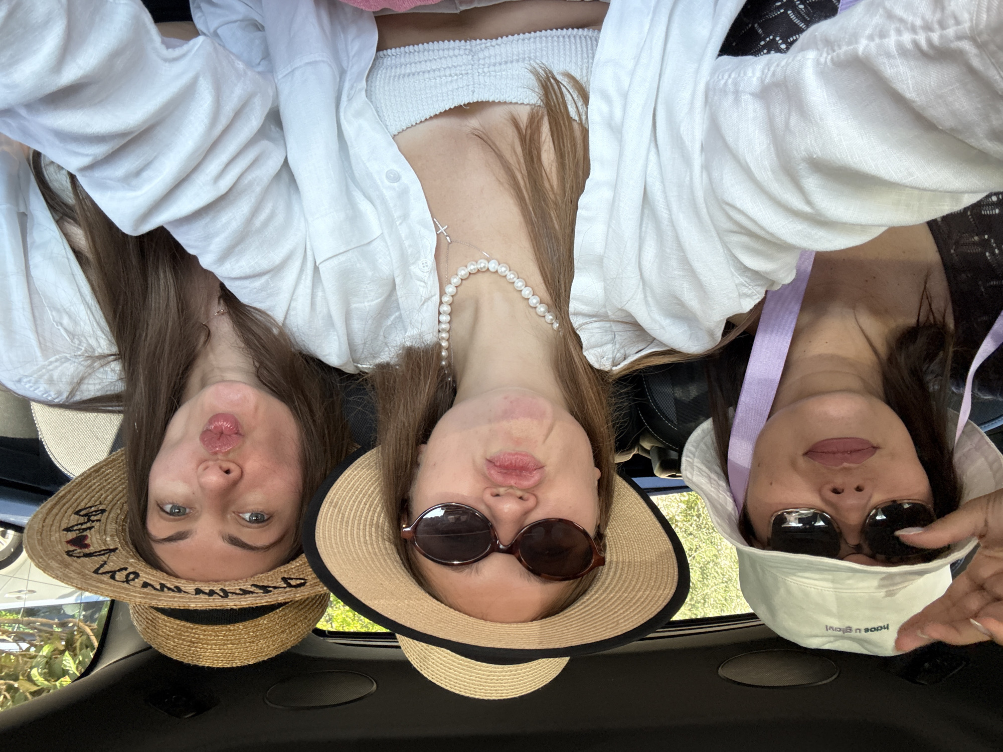 Three women are taking a selfie in a car. The woman on the left is wearing a white bucket hat with a purple ribbon and sunglasses. The woman in the middle is wearing a straw hat with a black band and sunglasses. The woman on the right is wearing a straw hat with a black band. All three women are wearing white tops and have their lips pursed. The car appears to be moving, as indicated by the greenery outside the window. The scene is well-lit, suggesting it is daytime. Based on the context provided, this image was taken in the city of Grad Šibenik, Croatia.
