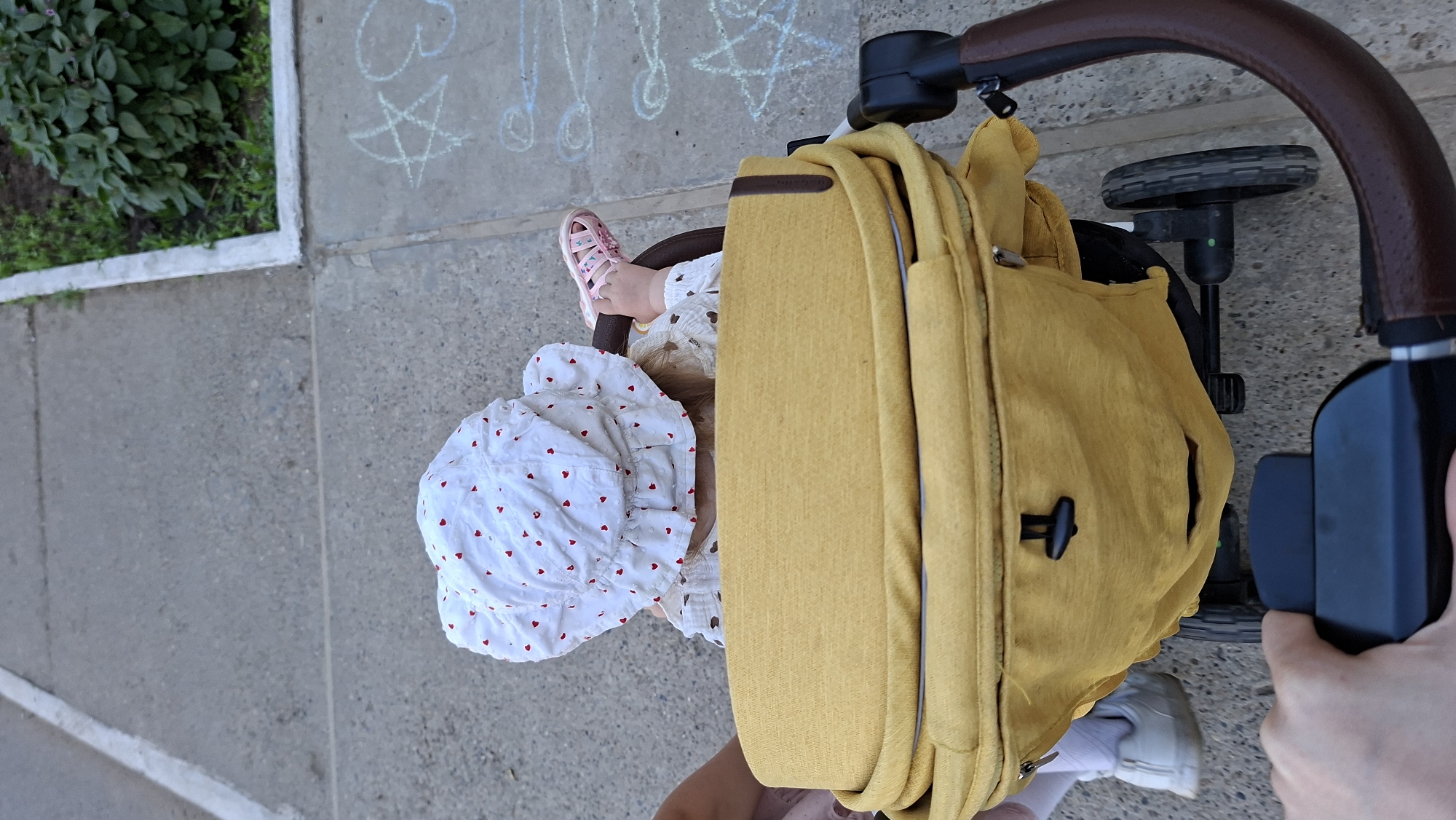 Captured from an elevated perspective, the image shows a young child in a stroller, being pushed along a concrete surface. The child is wearing a white bonnet adorned with red hearts, a patterned shirt, and pink sandals. The stroller is yellow and has a brown handlebar. A person's hand is holding the stroller's handlebar. The background includes chalk drawings on the concrete, a small patch of greenery, and a portion of a shoe. The location is Chaikovsky, Russia.