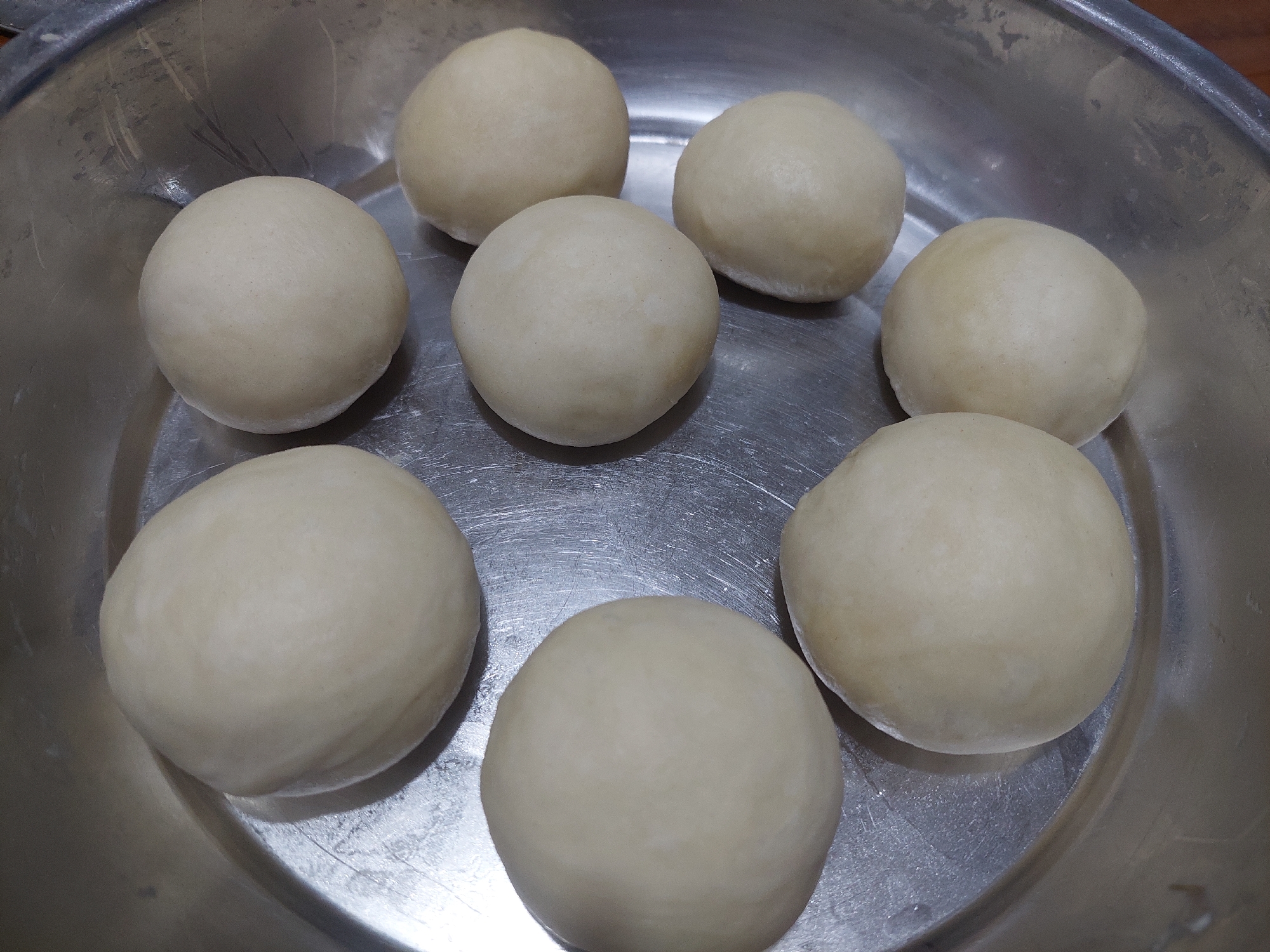 The image shows a close-up, overhead view of a metal bowl filled with eight pale, round balls of dough arranged in a circular pattern. The dough balls vary slightly in size and are uniformly light in color with a smooth texture. The metal bowl is shallow and appears to be made of stainless steel, exhibiting some minor scratches and reflections. The background is slightly out of focus, hinting at a wooden surface. The scene is static and focuses solely on the prepared dough. There are no people, animals, or other objects visible. The lighting is even, casting soft shadows from the dough balls onto the bowl. The image depicts a culinary preparation stage, likely for baking or cooking. Based on the provided context, this scene is located in the city of Jelutung, Indonesia.