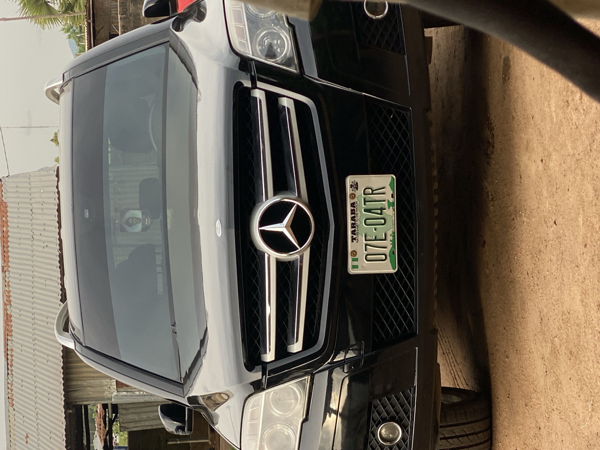 A black Mercedes-Benz GLK-Class SUV is parked on a dirt road in front of a building with a corrugated metal roof. The car's license plate reads "07E-04TR". A small framed picture of a person is hanging from the rearview mirror. The environment appears to be a rural or semi-urban area in Jalingo, Nigeria.