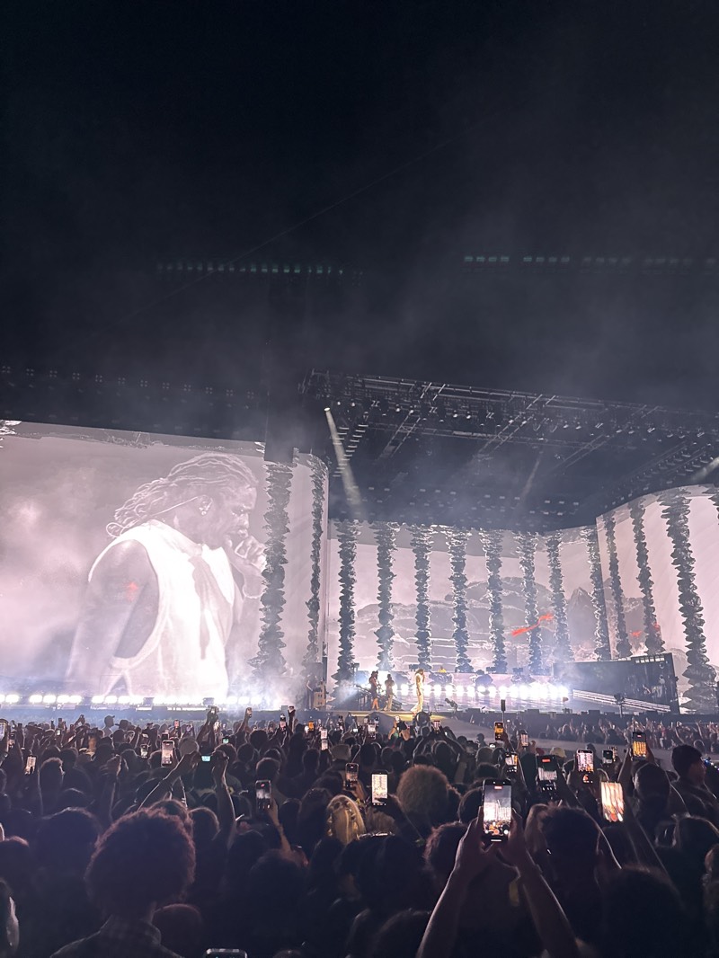 The image depicts a large crowd of people at a concert in Indio, California, United States. Many attendees are holding up their phones to record the performance on stage. A large screen to the left displays a black-and-white image of a performer. The stage has a complex structure with lights and other screens. The atmosphere appears lively and filled with energy.