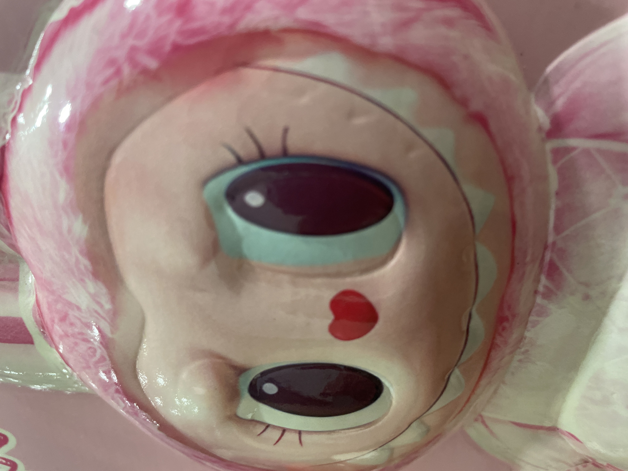 A close-up, inverted image of a pink-hued doll's face, possibly ceramic, is shown. The doll has large, dark eyes outlined in light blue, a small red heart painted on its cheek, and a pink and white striped headdress or bonnet. The scene lacks contextual information about a city or specific location.