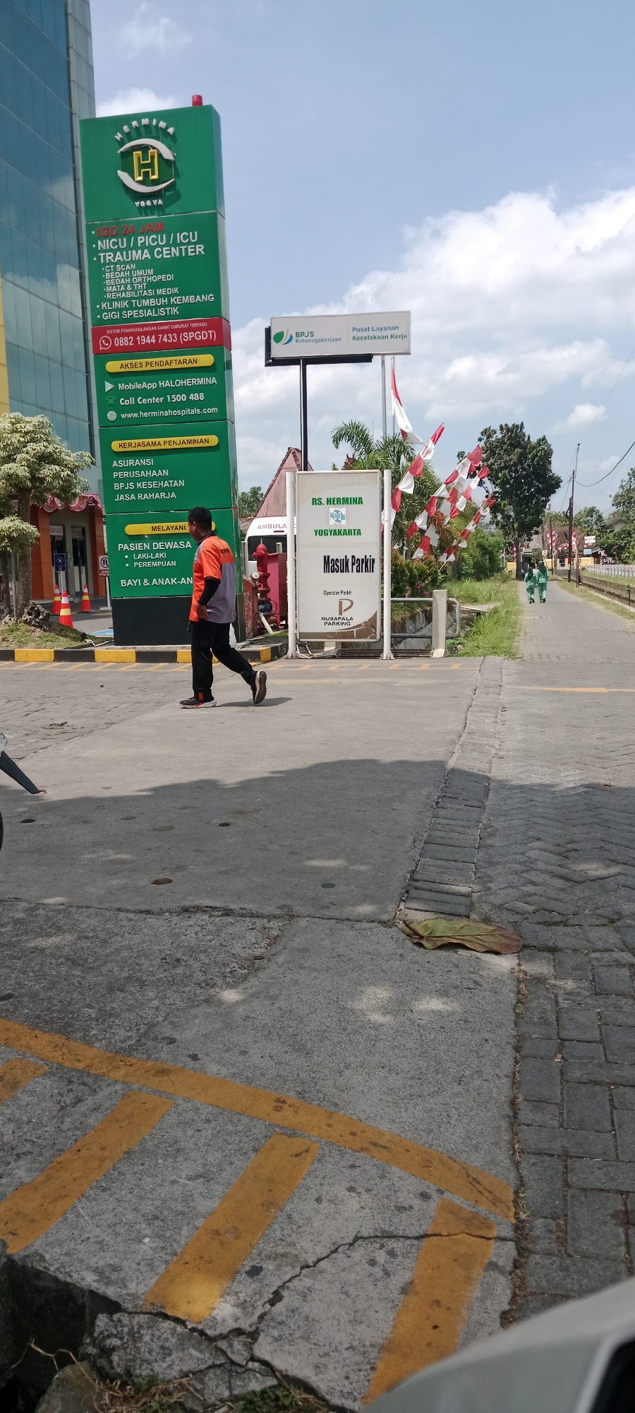 In Maguwoharjo, Indonesia, a man in an orange and gray uniform walks on a paved road in front of a hospital. The Hermina Yogyakarta Hospital's tall, glass-fronted building is visible on the left, with a green vertical sign listing hospital services like IGD 24 JAM, NICU/PICU/ICU, and TRAUMA CENTER. To the right of the hospital sign is another sign for RS. HERMINA YOGYAKARTA indicating "Masuk Parkir" (Enter Parking) with the Nusapala Parking logo. A BPJS Ketenagakerjaan sign, which translates to Social Security Agency for Employment, is also visible. Red and white Indonesian flags flutter on poles in the background. The road has yellow lane markings, and further down, two individuals in green uniforms are walking away from the camera. The sky is partly cloudy.