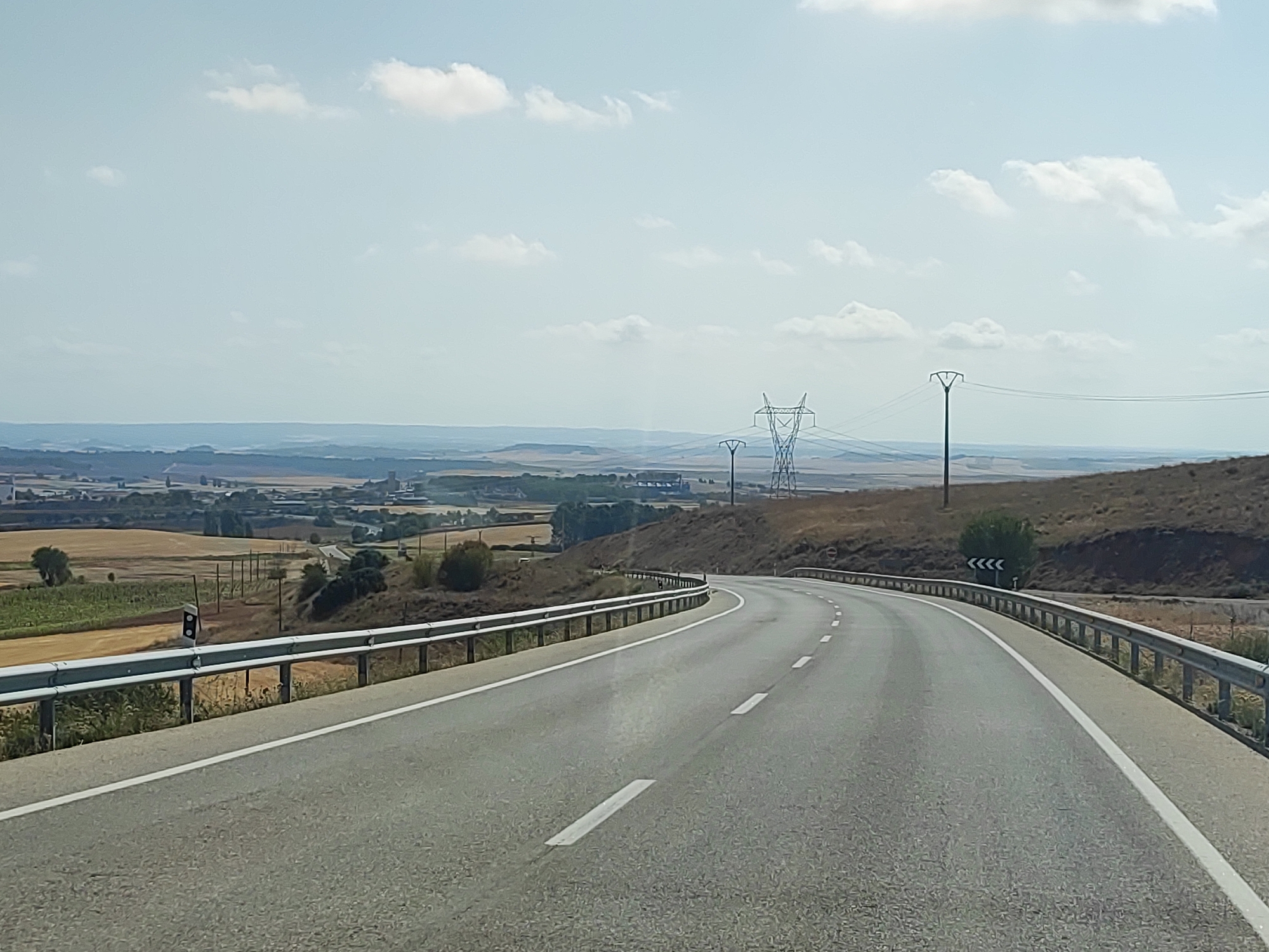 The image shows a winding asphalt road with white lane markings, curving towards the right, bordered by a metal guardrail on the left and a dry, grassy hillside on the right. In the background, a vast, hazy landscape unfolds, featuring agricultural fields, scattered trees, and distant buildings. Several power lines and tall, lattice-like transmission towers punctuate the horizon. The sky is a pale blue with scattered, wispy white clouds. The overall impression is of a rural road in a relatively arid environment under a clear sky. The location is identified as Castillo de Garcimuñoz, Spain.