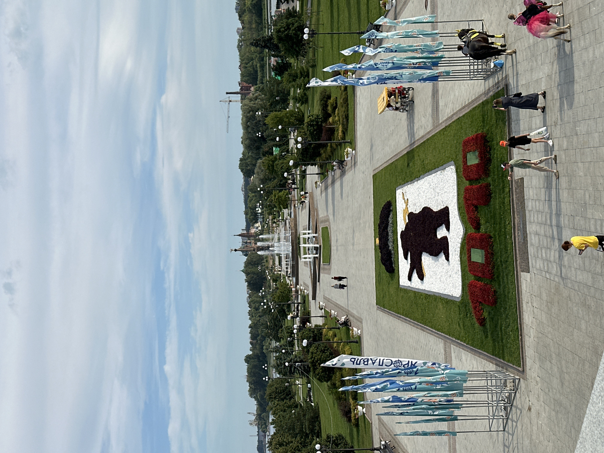 An aerial view captures a park scene in the city of Zhukovo, Russia. In the foreground, a large floral arrangement depicts the number "70" in red flowers, with a silhouette of a bear in a white floral background above it. A line of blue and white flags, some with Cyrillic text, stands to the left of the floral display. To the right, a black horse adorned with yellow leg coverings stands near a metal fence, while a small pony dressed in a pink tutu is also present. Several individuals are walking across the paved area: two adults and a child are walking away from the viewer, a woman in a black dress is taking a photograph, and a person in a yellow shirt is looking down at their phone. Further back, a pedestrian walkway extends into the distance, lined with manicured green lawns, trees, and lampposts. A large fountain with multiple tiers is visible in the middle ground. The background features more trees and buildings, with a construction crane and industrial structures on the horizon. The sky is partly cloudy.