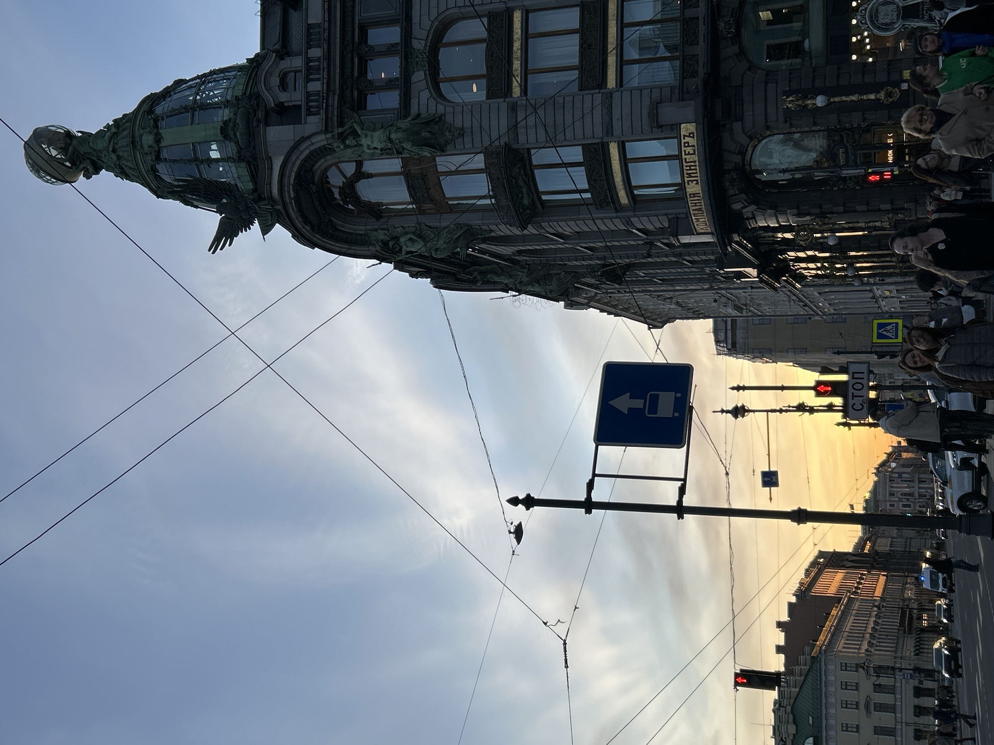 The image depicts an urban street scene in Saint Petersburg, Russia, captured during what appears to be sunset or late afternoon, given the warm, golden light illuminating the horizon.
The most prominent subject is the ornate building on the left, identified as the Singer House (Dom Knigi). This Art Nouveau structure features intricate architectural details, including sculpted eagles and decorative elements on its facade and windows. Its distinctive glass-domed tower, topped with a large glass globe, stands out against the sky. Vertically oriented Cyrillic text on the building reads "КОМПАНИЯ ЗИНГЕРЪ" (KOMPANIYA ZINGER'), translating to "Singer Company".
Below the building, a busy street scene unfolds with pedestrians walking on the sidewalk and what appear to be parked or moving vehicles on the road. The sky transitions from a light blue at the top to a vibrant golden hue towards the lower right, where distant buildings are bathed in the warm light.
Numerous elements of street furniture are visible, including poles, overhead wires (likely for trolleybuses), and traffic lights, some displaying red signals. A blue road sign with a white arrow and a bus icon points to the left, indicating a directional route or bus stop. Another smaller blue sign below it also shows a bus stop symbol. On a pole near a traffic light, the Cyrillic word "СТОП" (STOP) is clearly legible. A yellow pedestrian crossing sign is also visible on the right side of the street. The scene conveys a bustling city environment during the golden hour.