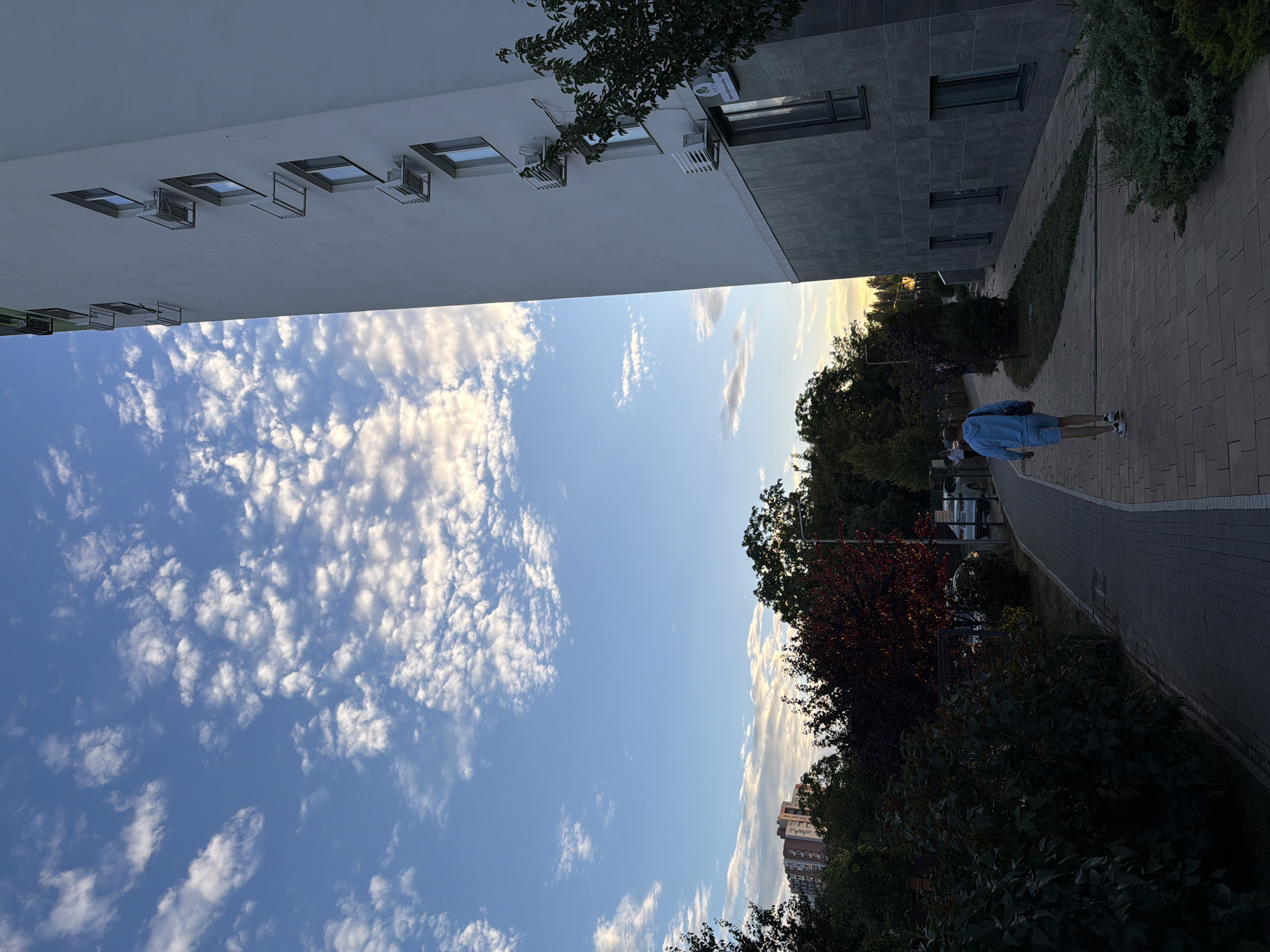 A person walks away from the camera on a paved path next to a large, modern building with gray stone accents and multiple windows. The building is tall and white, with several air conditioning units mounted on its exterior. The sky is blue with scattered white clouds, suggesting it is daytime. There are trees and other foliage visible in the background, as well as a distant apartment building. The overall scene appears to be in an urban or suburban setting.