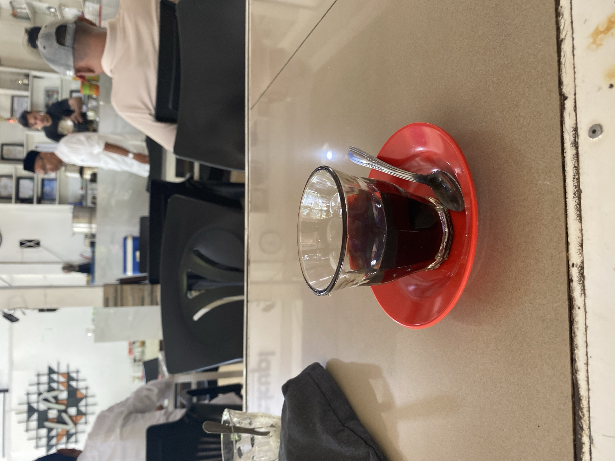 A low-angle shot captures a glass of dark liquid, likely coffee, resting on a red saucer on a beige table. A spoon is placed on the saucer. To the left, a crumpled dark fabric item is visible. In the background, several people are seated at tables, and others are standing behind a counter in what appears to be a cafe. A decorative wall art depicting geometric patterns is visible on the left wall. The cafe appears to be in Lhokseumawe, Indonesia, based on the wall art which has "LSM" in the center, a common abbreviation for Lhokseumawe.