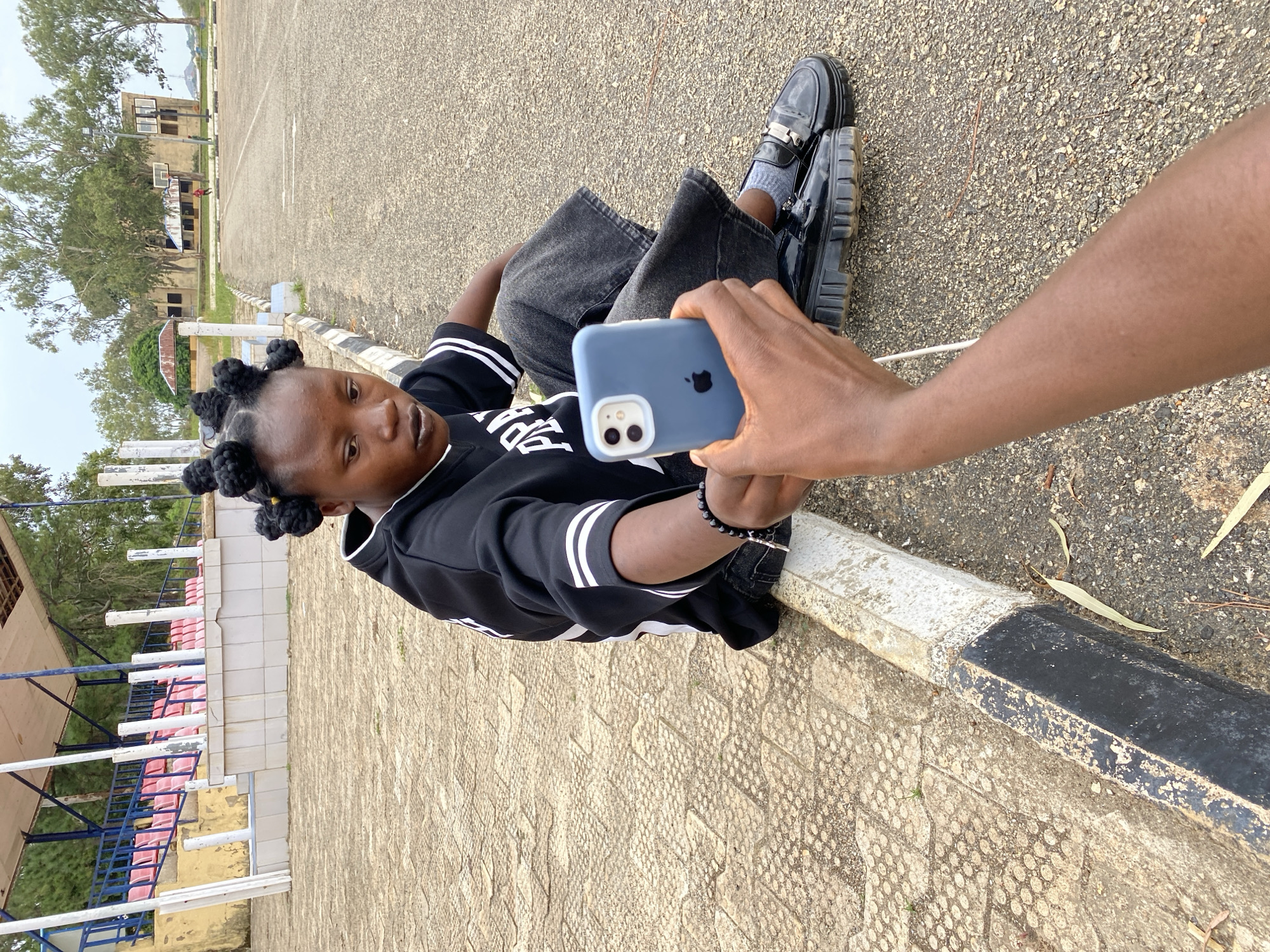 A young Black woman with braided hair sits on the edge of a paved area next to a curb in Zawan, Nigeria. She is looking down at a blue smartphone held by an unseen person. The woman is wearing a black hoodie with white stripes on the sleeves and dark pants. She has a black beaded bracelet on her left wrist. The background shows a stadium with red seats, trees, and buildings, suggesting an outdoor public space such as a school or sports complex. The ground is a textured paving stone near the curb, transitioning to asphalt further away. The lighting indicates it is daytime, possibly overcast.
