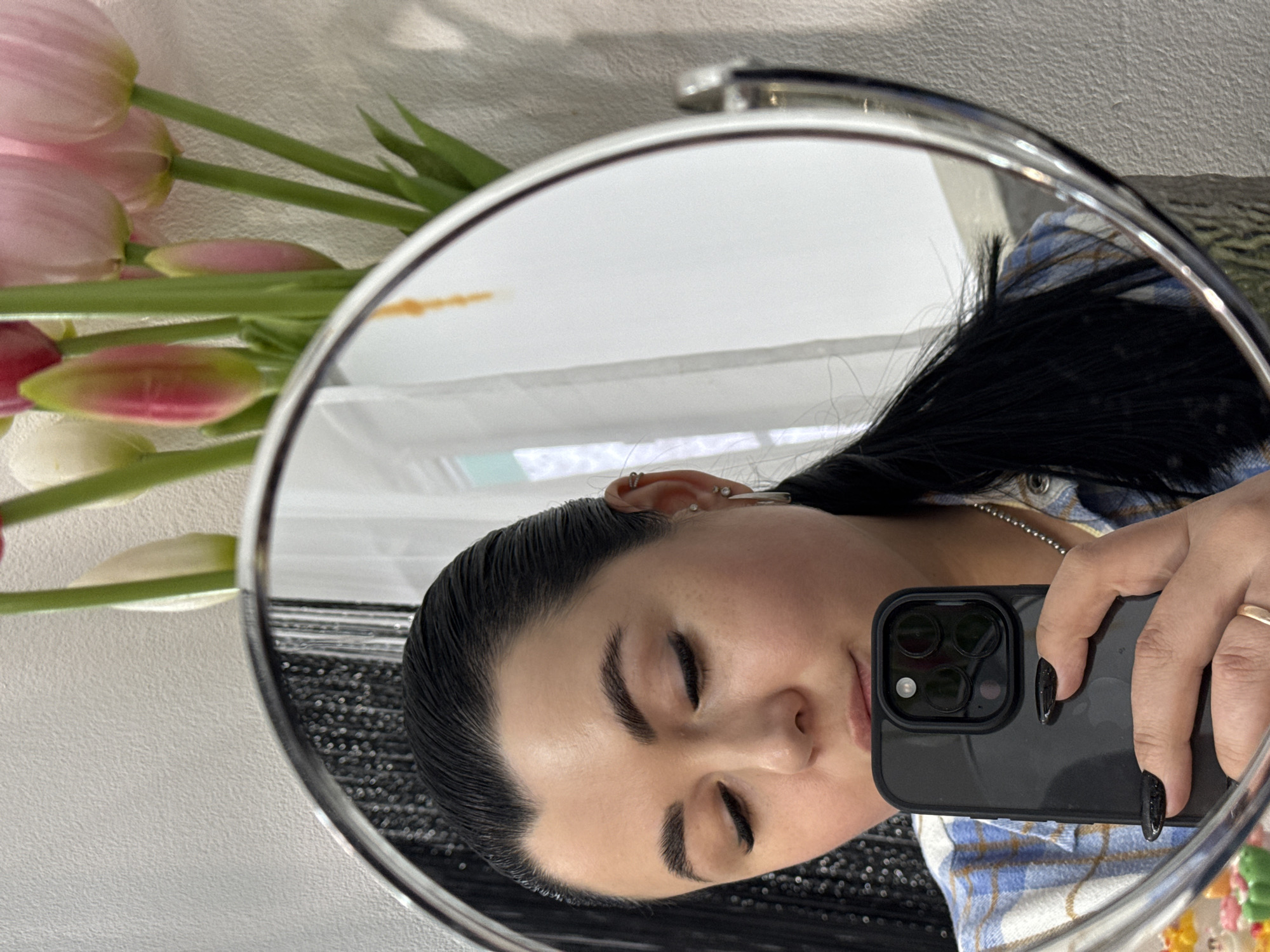 A woman is reflected in a round mirror. She is holding a smartphone to take a selfie. She has dark hair and is wearing a plaid shirt. A bouquet of pink tulips is visible near the mirror. The location is implied to be Chaikovsky, Russia.