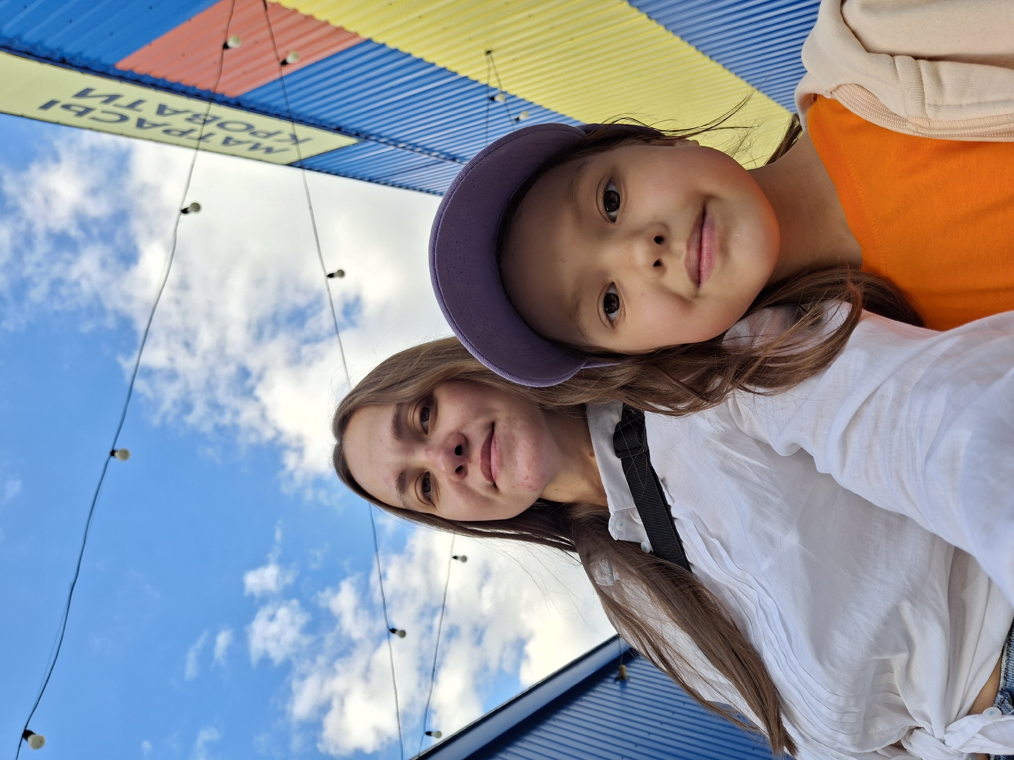In the city of Chaikovsky, Russia, two individuals, an adult female and a young girl, are captured in a selfie-style photograph against a backdrop of a bright blue sky with scattered clouds and a vibrantly colored corrugated metal structure. The adult, positioned on the left, has long brown hair and is wearing a white collared shirt tied at the waist. The young girl, on the right, sports a purple baseball cap and an orange t-shirt, layered with a light-colored zip-up hoodie. They are standing in an outdoor setting, possibly an alley or courtyard, with strings of light bulbs strung overhead. The corrugated structure features panels of blue, yellow, and red, with Russian text that translates to "MATRASSY KROVATI," indicating a store selling mattresses and beds. The lighting suggests it is daytime.