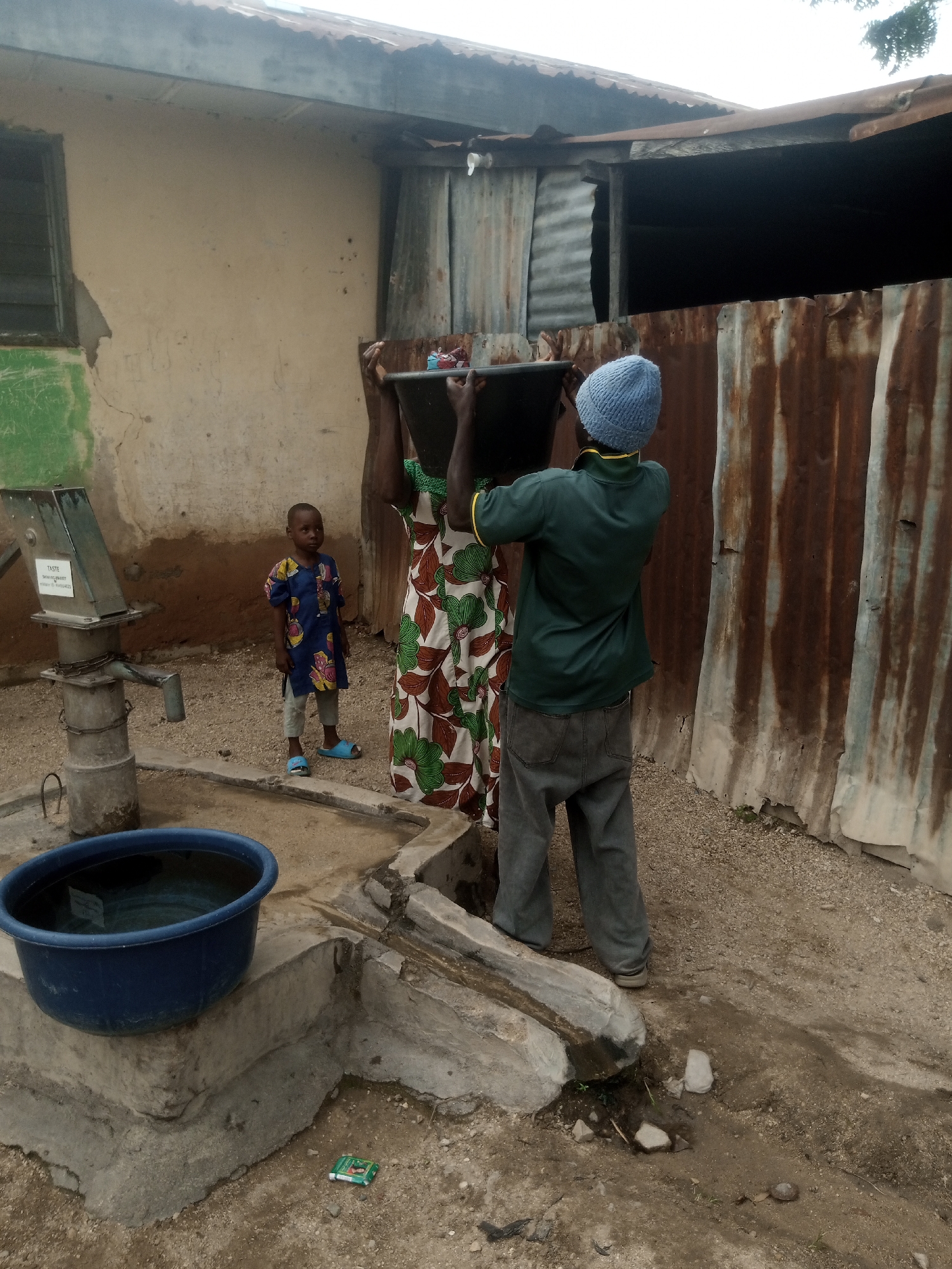 An eye-level, medium shot captures a rural outdoor scene in an undetermined location. In the foreground, a large blue basin filled with water rests on a concrete structure. To the left, a manual water pump stands tall, its metal components weathered and aged. A young boy in a blue and yellow patterned dress and blue sandals stands near the pump, looking towards the right. In the center, a person wearing a patterned dress and a blue knitted cap is holding a large black basin over their head, seemingly assisting another person with the basin, whose arms are also visible. The background is dominated by a rustic structure with corrugated metal walls and a tin roof, suggesting a modest dwelling or outbuilding. The ground is covered with gravel and dirt.