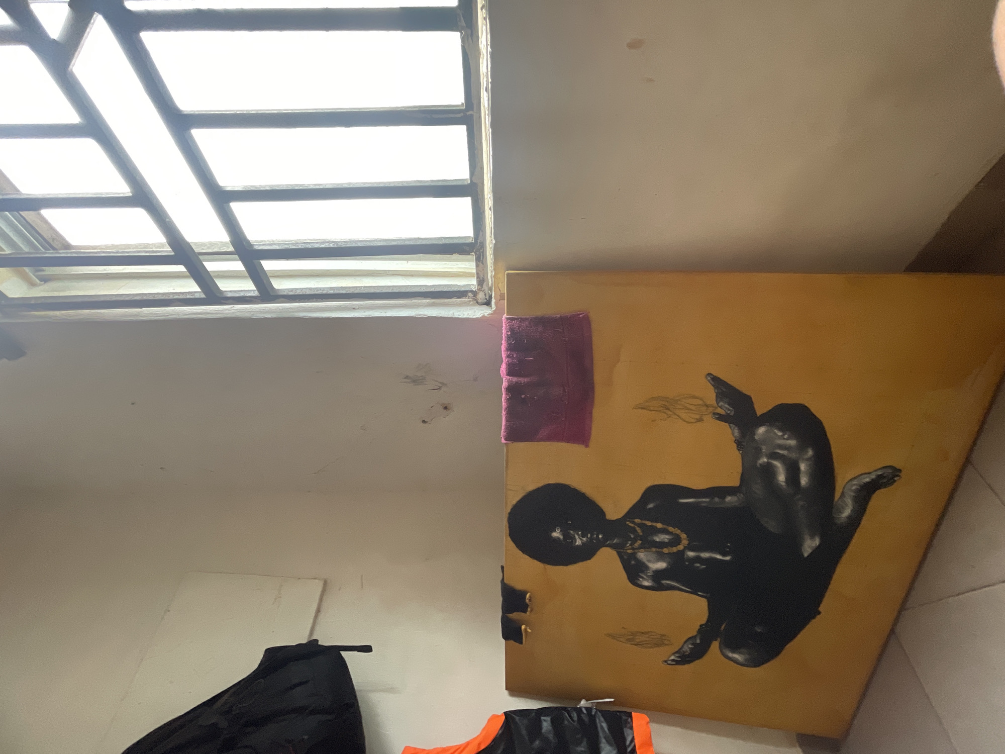 The image portrays an indoor scene, likely a room in Ado, Nigeria, illuminated by natural light entering through a window. The overall ambiance suggests a casual or functional space.
In the upper left quadrant, a high, barred window provides the primary light source, indicating daytime with a bright, possibly clear, sky outside. The window features dark, gridded metal bars, and its frame and the surrounding wall show signs of age or wear.
Dominating the right side of the frame is a large, rectangular painting, mounted or leaning against a wall. The artwork features a stylized, dark-skinned human figure with a proportionally large head and a smaller body, depicted in a reclining or floating posture. The figure is adorned with a gold-colored beaded necklace, and abstract, wispy shapes surround it. The background of the painting is a solid mustard yellow or light brown. A small, dark magenta cloth is visible, tucked into the top edge of the painting.
The walls are painted in a light cream or off-white color, exhibiting minor marks and inconsistencies. Towards the bottom of the image, sections of what appear to be clothing or bags are visible: a black item on the bottom left and an orange and black item towards the bottom right. A small portion of light-colored tiled flooring is also visible in the bottom right corner. No specific activity is occurring, and there is no visible text in the image.