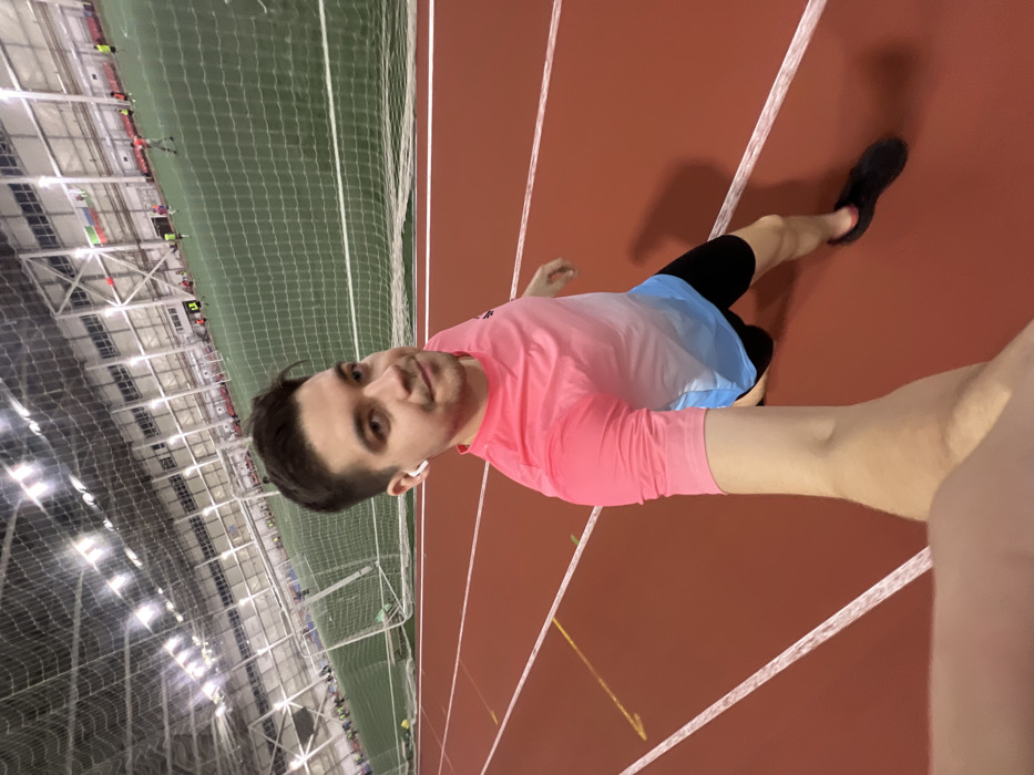 The image shows a young man posing for a selfie on an indoor running track. The photo is rotated 90 degrees clockwise, so viewed upright, the man is standing on a red track with white lane markings. He is positioned slightly off-center, facing the camera with a neutral expression and a faint smile.
The man has short, dark hair, neatly styled, and prominent dark eyebrows. He is wearing a short-sleeved athletic shirt that transitions from bright pink on top to light blue at the bottom. He also wears black athletic shorts and a black athletic shoe on his right foot, which is slightly raised as if in mid-step or preparing to move. A white wireless earbud is visible in his right ear. His right arm is extended into the foreground from the right edge of the frame, presumably holding the camera, while his left arm is bent at the elbow.
The background reveals a spacious indoor sports facility. Immediately behind the man and the running track is a tall green netting, likely separating the track from a larger sports field, possibly for soccer or another indoor sport. Beyond the netting, the facility features a high ceiling with numerous artificial lights and intricate metal support beams, indicative of a large modern arena or gym. Small, indistinct figures of other people can be seen in the far background on the green field, suggesting an ongoing activity or training session. The overall lighting is bright and even, consistent with an indoor environment.