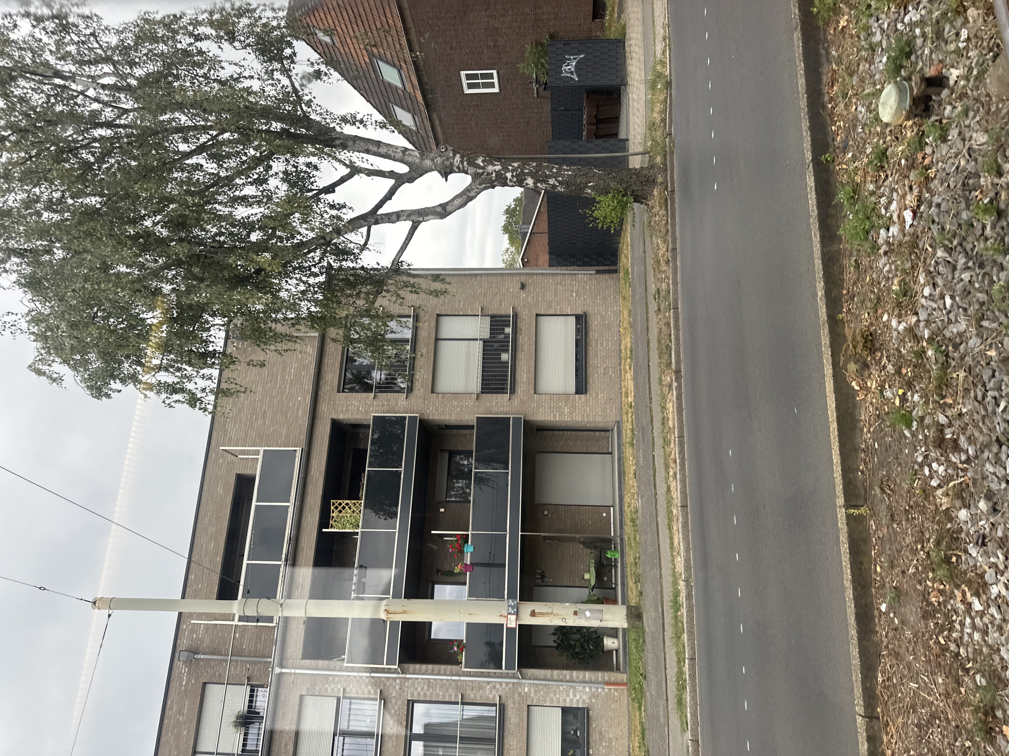 The image depicts a street scene in Krefeld, Germany. In the foreground, a paved road with white dashed lane markings runs horizontally. The roadside is lined with a concrete curb and a strip of gravel and sparse vegetation. Beyond the road, a multi-story apartment building with a brick facade and balconies with black glass railings is visible. A tall, slender utility pole stands in front of the building, with overhead wires stretching across the sky. To the right of the apartment building, a red-brick house with a tiled roof is situated, featuring a small white window. A large deciduous tree with a textured, light-colored trunk and abundant green foliage stands between the apartment building and the red-brick house. Behind the tree and fence, brown waste containers are partially visible. The sky is overcast, suggesting a cloudy day.