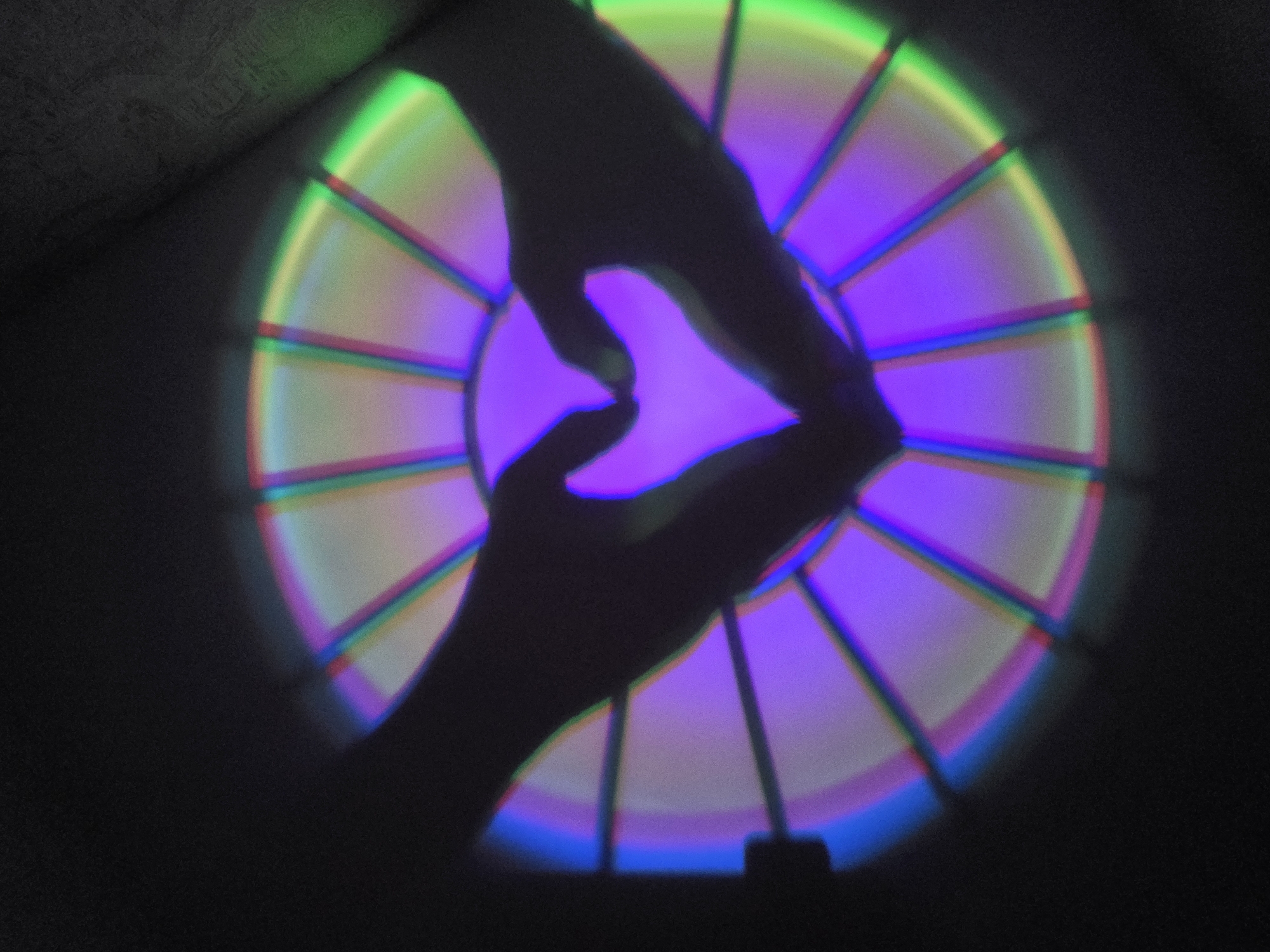 The image shows a dark setting with a bright, circular light pattern projected onto a surface. The light pattern is divided into segments, each illuminated with different colors including green, red, blue, and purple. In front of the light pattern are two shadowy hands forming a heart shape. The hands are dark silhouettes against the bright light. The lower edge of the circular light pattern reveals the projector. There is no information to suggest the image was taken in the city of Chaikovsky, Russia.