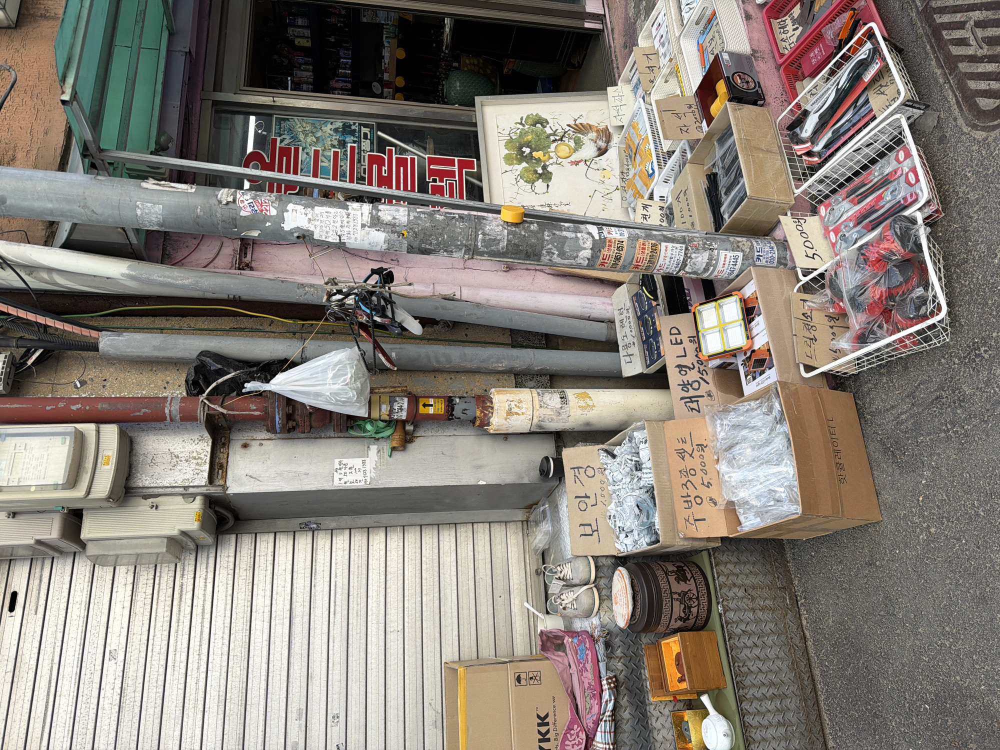 The image presents a cluttered street scene in Seoul, South Korea. An assortment of items for sale is displayed along the sidewalk in front of a storefront. These items include tools, hardware, safety glasses, and household goods arranged in baskets and cardboard boxes. There are power meters and electrical wiring along the wall above a closed metal shutter. Korean signage is visible on the storefront and on price tags throughout the display. The overall impression is of a small business extending its sales area onto the sidewalk.