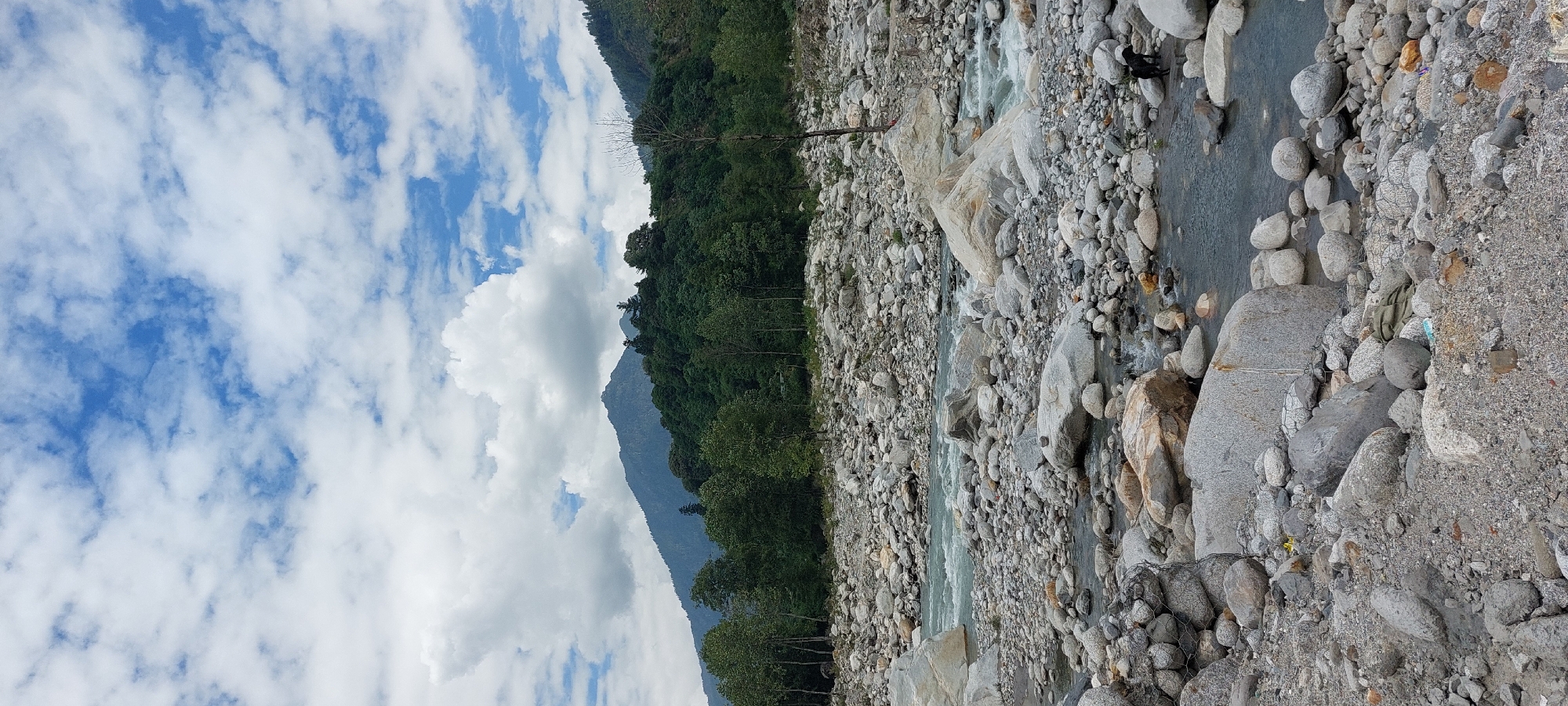 The image depicts a rocky riverbed in Kalath, India, with a flowing river and lush green mountains in the background. The sky is partly cloudy, with white clouds against a blue sky. The foreground is dominated by numerous gray and white stones of varying sizes, some partially submerged in the water. A small black dog is visible near the river's edge on the right side of the frame. The river itself is clear and shows signs of movement, with ripples and small rapids. In the middle ground, the river flows between larger rocks, and further back, dense green trees cover the lower slopes of the mountains. The mountains rise steeply behind the trees, with their peaks reaching towards the sky. The overall scene conveys a natural and serene environment.