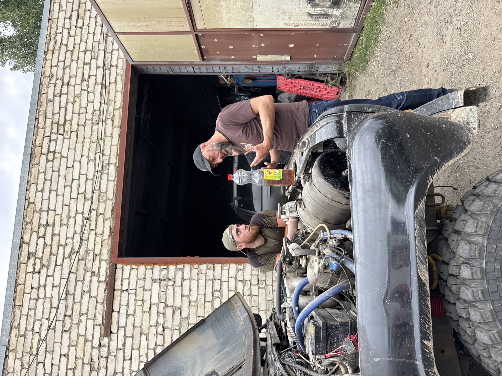 Two men are standing in front of an open-hooded black off-road vehicle parked in front of a brick garage. The man on the left, wearing a green cap and a dark olive t-shirt, is leaning over the engine. The man on the right, wearing a brown t-shirt and blue jeans, is holding a plastic bottle filled with amber liquid and gesturing with his hands. The garage appears to be in a rural or industrial area, with gravel on the ground and a large metal door. A tree is visible in the background. The image is likely taken in Karachayevsk, Russia.