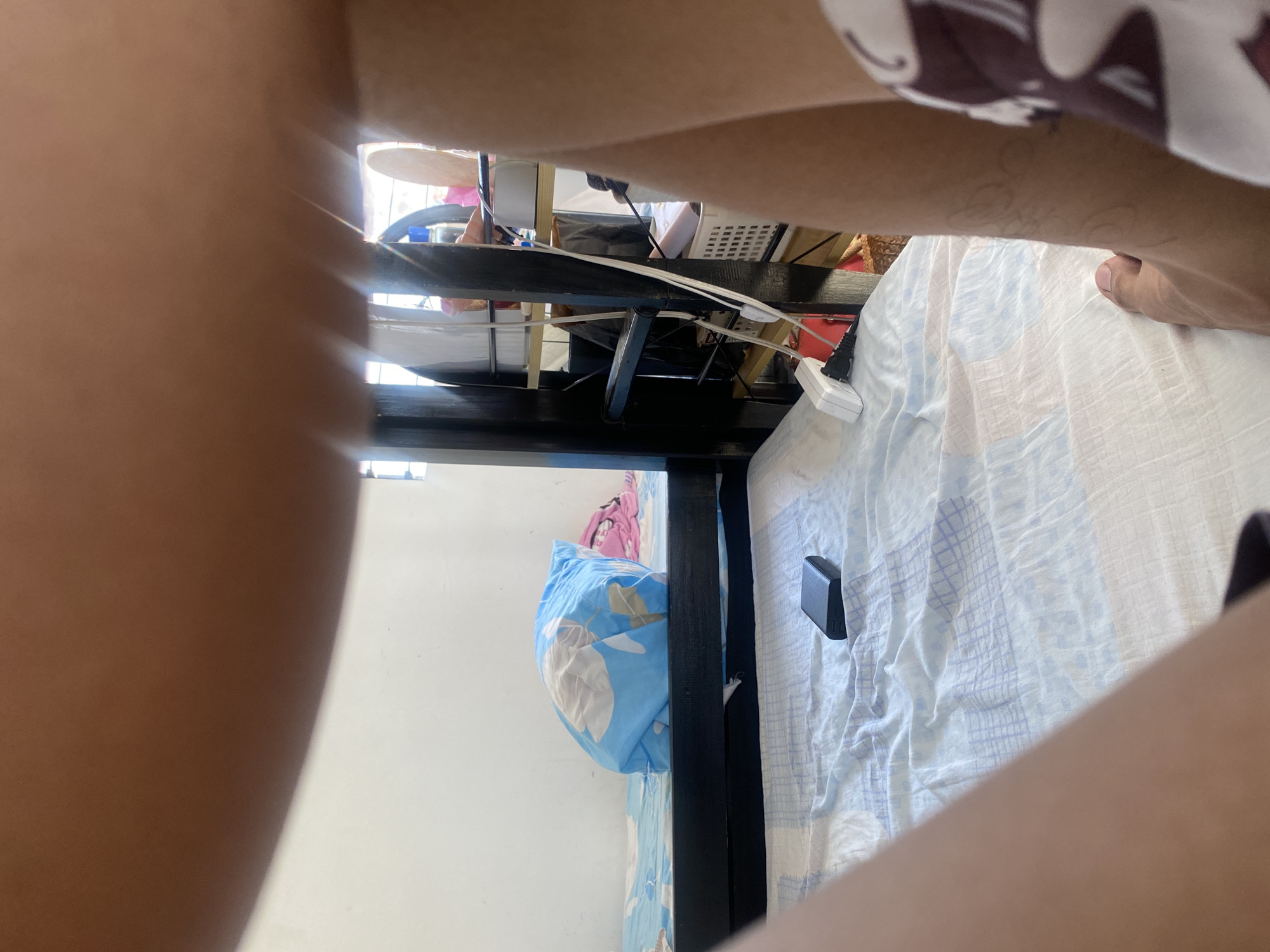 This image, rotated 90 degrees clockwise, captures an indoor scene, likely a bedroom, from a low and upward-angled perspective. Large, blurred sections of human skin, possibly arms or legs, frame the immediate foreground on the left and right, with a bare foot visible on the top right (when correctly oriented).
The central area features a bed covered with a wrinkled, light-colored sheet, which appears to be white with faint blue grid-like patterns. Resting on the sheet is a small, black rectangular object, possibly a mobile phone or power bank. A blue pillow with a white floral or leafy design is positioned near the bed frame, with a pink fabric item behind it. A dark, sturdy bed frame, possibly made of wood, runs along the left side of the bed.
Beyond the bed, the background reveals a cluttered space, possibly a desk or storage area. Bright sunlight illuminates this section from an unseen window or opening on the top left. Visible objects include a light-colored circular item (possibly a table surface or part of a drum), a white power strip with several cables plugged in and hanging down, and other indistinct items such as a white plastic basket and dark bags. The scene depicts a casual, daytime setting within a personal living space. No visible text is present.