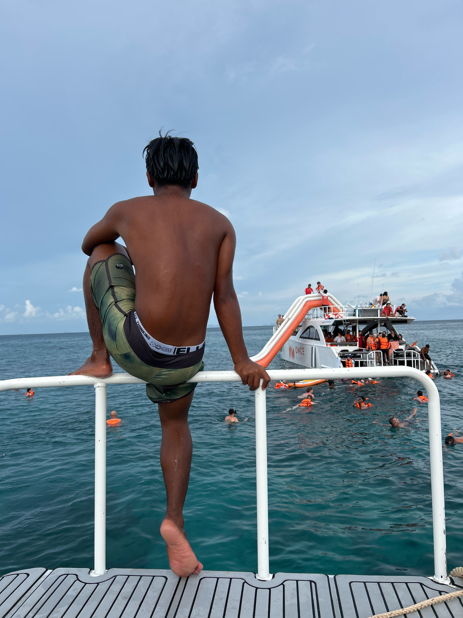 A young man with dark hair, wearing green and black patterned shorts, sits on a white railing, looking out at a boat filled with people in the ocean. The boat has an orange slide. Many people in the water are wearing orange life vests, suggesting they are swimming or snorkeling. The water is a clear, turquoise color. The sky is overcast. The scene appears to be at a tourist destination, possibly near Rawai, Thailand, given the presence of a large tour boat and people enjoying water activities.
