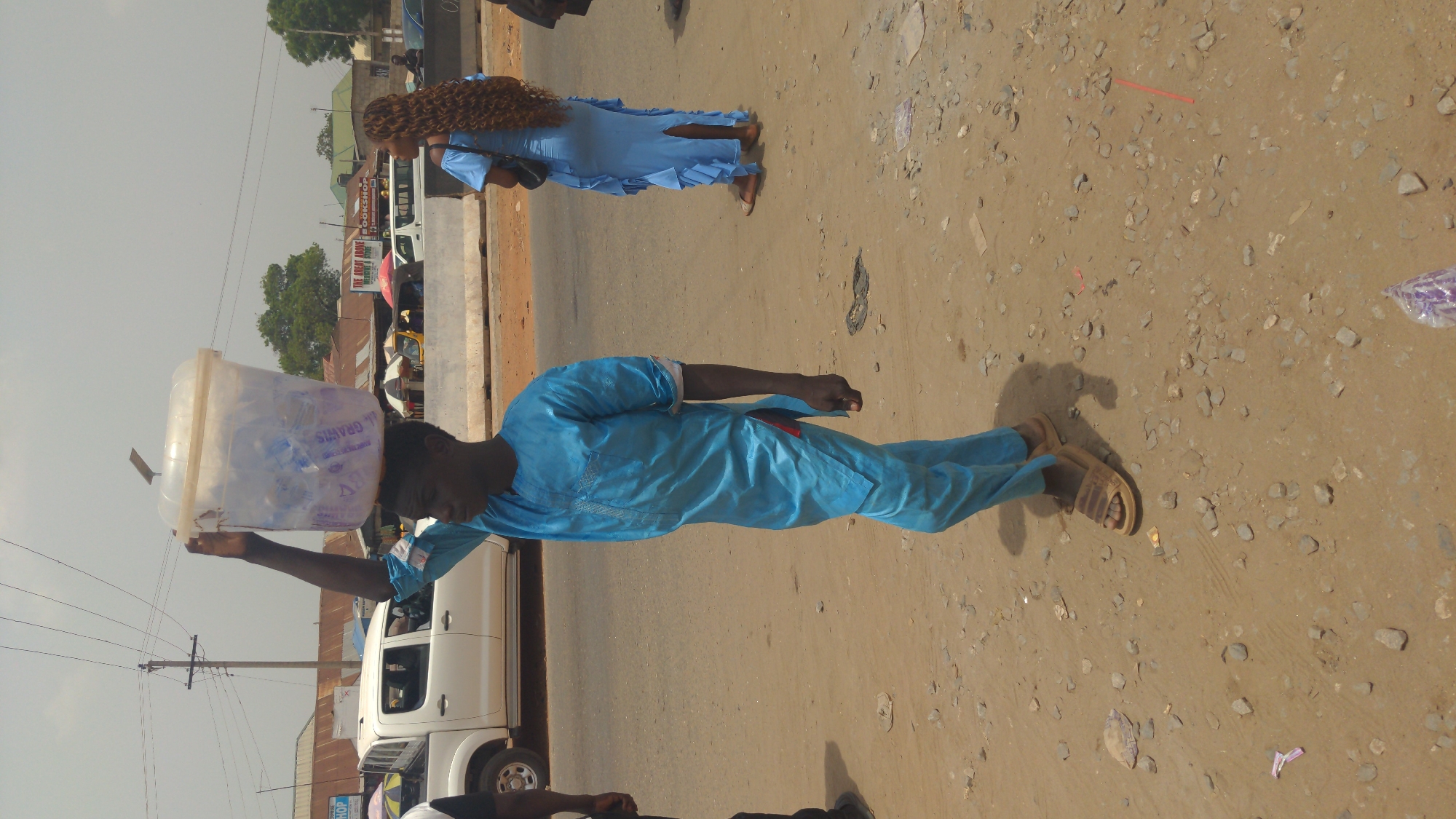 A man in a blue tunic and sandals balances a plastic bucket filled with merchandise on his head as he walks on a dirt and gravel shoulder next to a paved street in Ado, Nigeria. A woman in a similar blue dress with long curly hair walks ahead of him. A white pickup truck and other vehicles are parked on the street near storefronts. Utility poles with lines are visible.