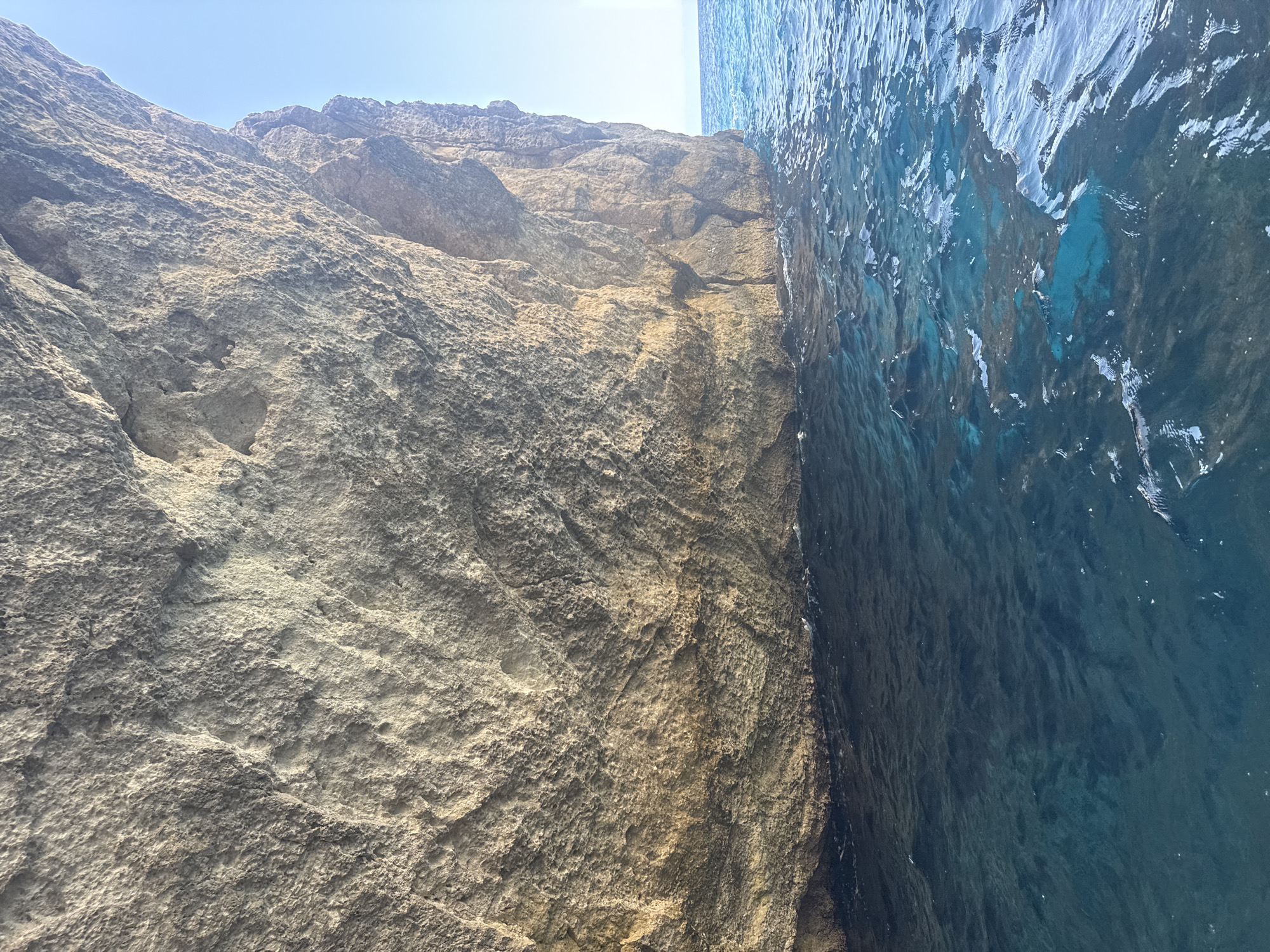A low-angle, wide shot captures the rough texture of a large, weathered rock face that dominates the left side of the frame. The rock is a mix of earthy brown and gray tones, showing pitting and irregularities from erosion. It extends from the bottom of the image upwards and to the right, creating an overhang. Beneath the rock face, the deep blue ocean fills the lower portion of the image. The water is choppy, with whitecaps indicating movement. Patches of lighter, turquoise water are visible beneath the surface, suggesting shallowness or submerged features. The sky above is a clear, pale blue, with the horizon line visible in the distance to the right. The scene is devoid of people or any man-made objects, suggesting a natural, uninhabited coastal environment.