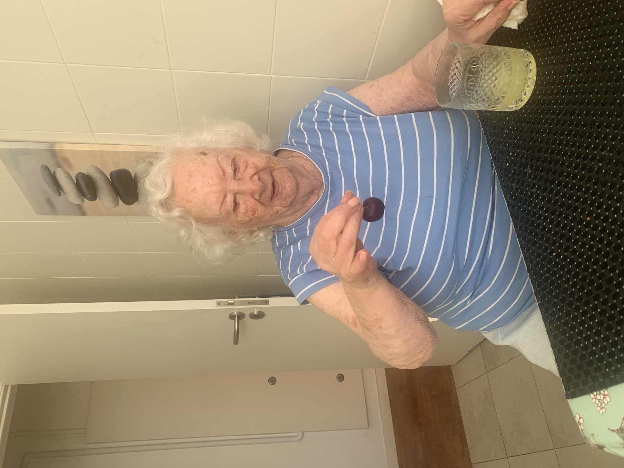 An elderly woman with white hair and fair skin, wearing a blue and white striped t-shirt, sits at a table holding a dark purple plum. A glass of yellow liquid, likely juice, is on the table in front of her, to her right. A white napkin is also visible on the table, partially obscured by her hand. The background consists of a tiled wall and a partially open door leading to another room. A framed picture of stacked stones hangs on the wall. The scene appears to be indoors, possibly in a kitchen or dining area. There are no visible indications of the location being in Lisbon, Portugal.