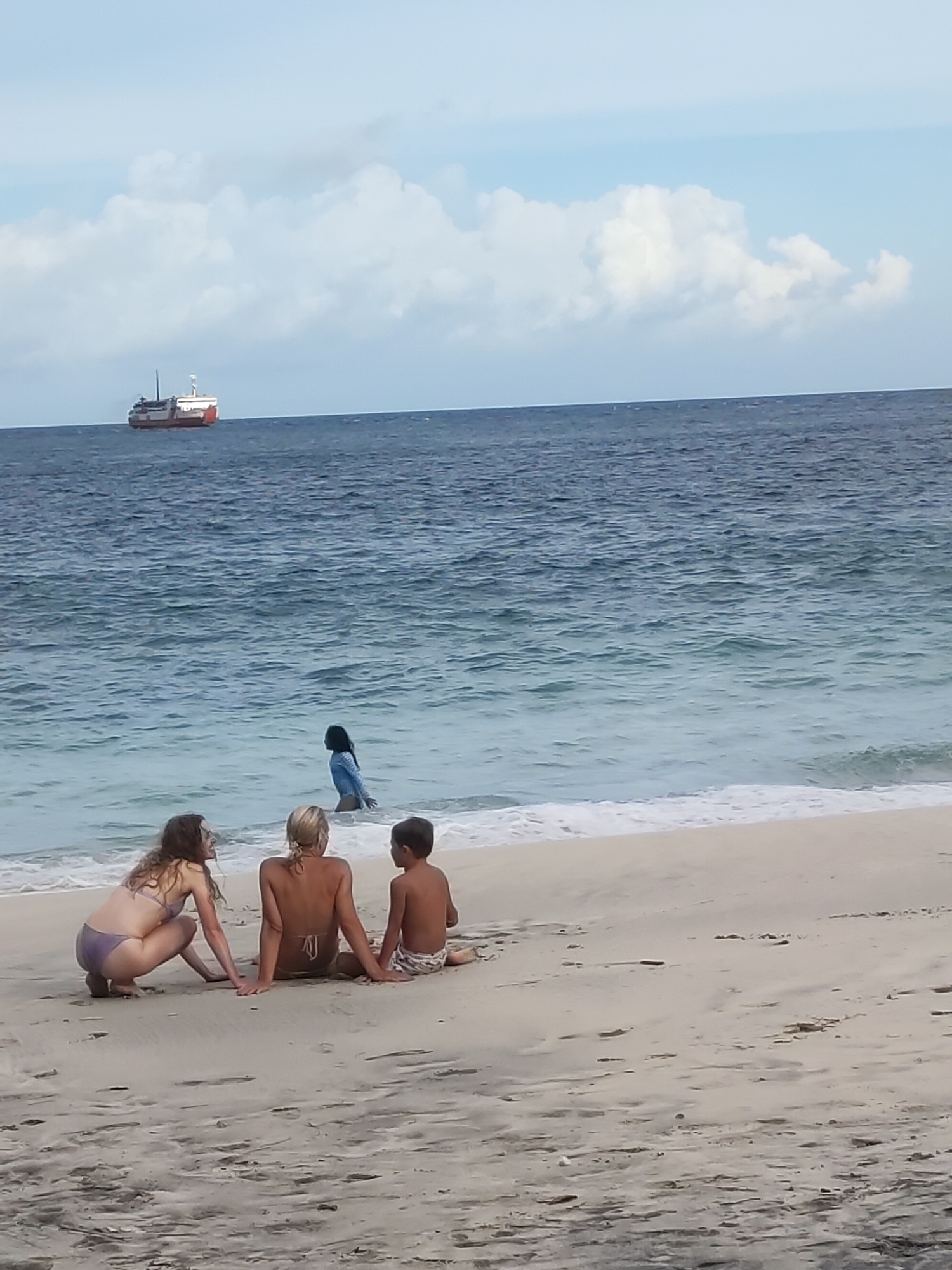 Padangbai, Indonesia: Two light-skinned women and a boy are seated on a sandy beach, facing the ocean. One woman is crouching while the other woman and boy are sitting. A person in blue swimwear is wading in the water. A large ship is visible in the distance. The sky is partly cloudy.