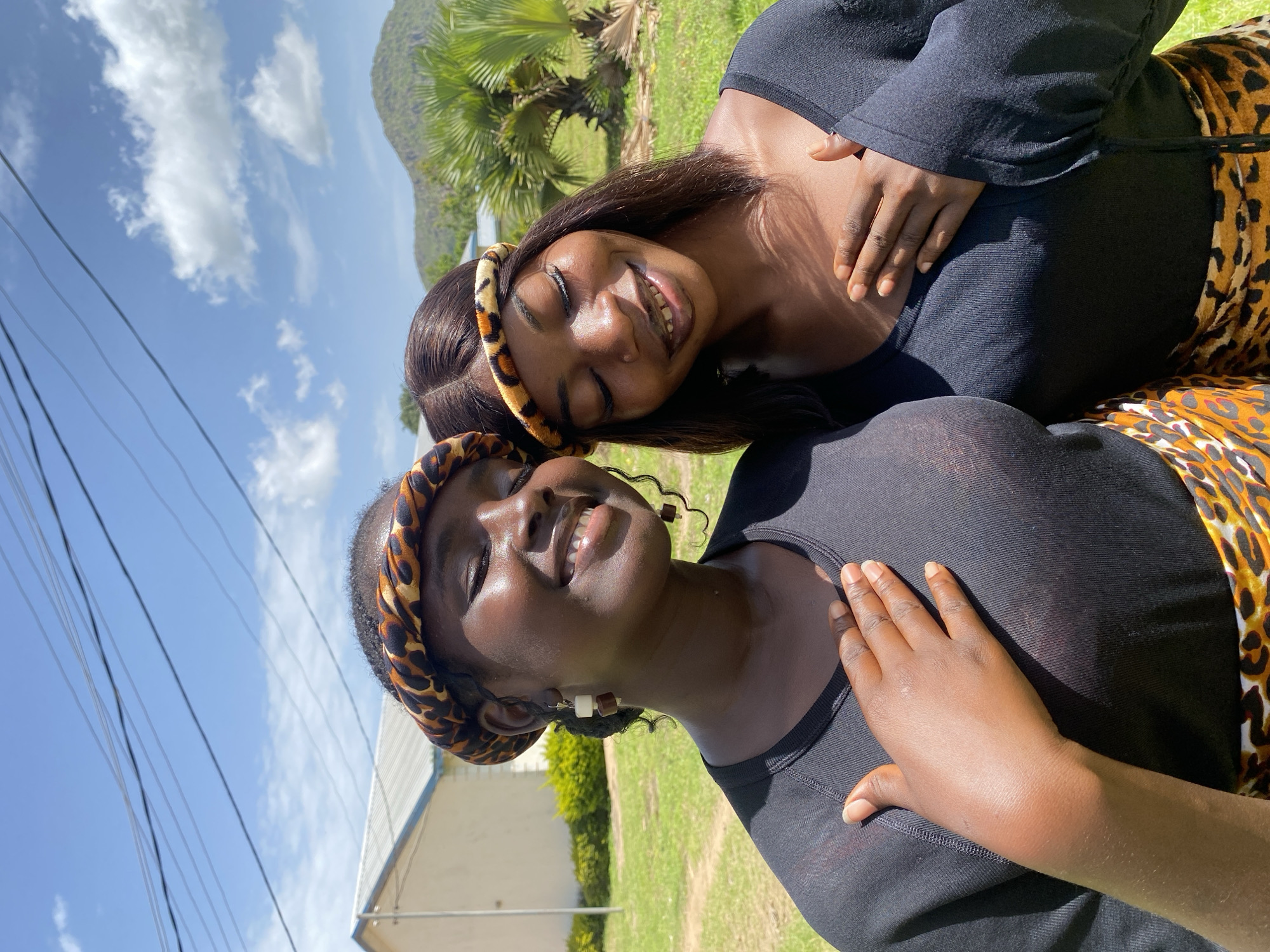 This media file captures a close-up of two young Black women outdoors, likely in Nigeria, as indicated by the location context. The image is oriented vertically, with the sky taking up a significant portion of the background. The sky is bright blue with scattered white clouds, suggesting a sunny day. In the distance, there are green hills and palm trees, further supporting an outdoor, possibly tropical or subtropical, setting. Power lines are visible in the upper left quadrant, and a building with a white roof and some green shrubbery are present in the lower portion of the image.
The two women are positioned in a way that suggests intimacy or close friendship. Both are wearing black tops and have their hair styled naturally. They are also adorned with leopard-print headbands. The woman in the foreground has her eyes closed and is smiling broadly, appearing relaxed and happy. Her hand is resting on her chest. The woman behind her is also smiling, with her eyes open, and her head is tilted towards the other woman. Her hand is resting on the shoulder of the woman in front. Their clothing includes black tops and leopard-print wraps or skirts. The overall mood is joyful and positive, evoking a sense of warmth and connection. The time of day appears to be daytime, given the bright sunlight and clear sky. There is no visible text in the image.