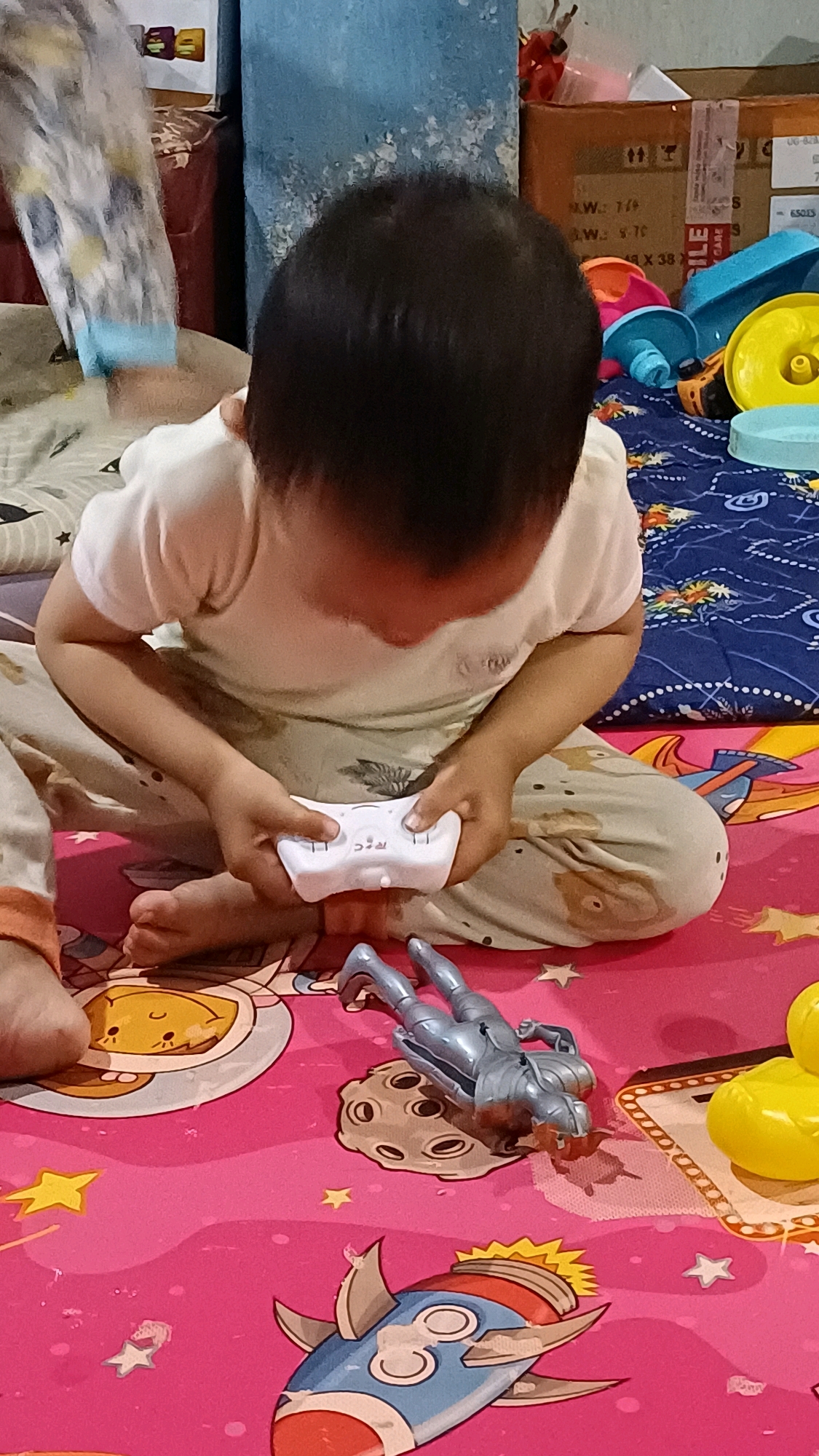 In Wonokerso, Indonesia, a young child is seated on a pink mat adorned with a space-themed pattern, including rockets, stars, and planets. The child is wearing a white t-shirt and light-colored pants with a subtle print. They are holding a white remote control, with their fingers positioned over buttons, suggesting engagement with a toy. To the right of the child, a silver robot action figure lies on its back on a depiction of a moon. Further to the right, a yellow toy, possibly a duck, is partially visible. In the background, there are cardboard boxes stacked and a blue wall with a textured surface.