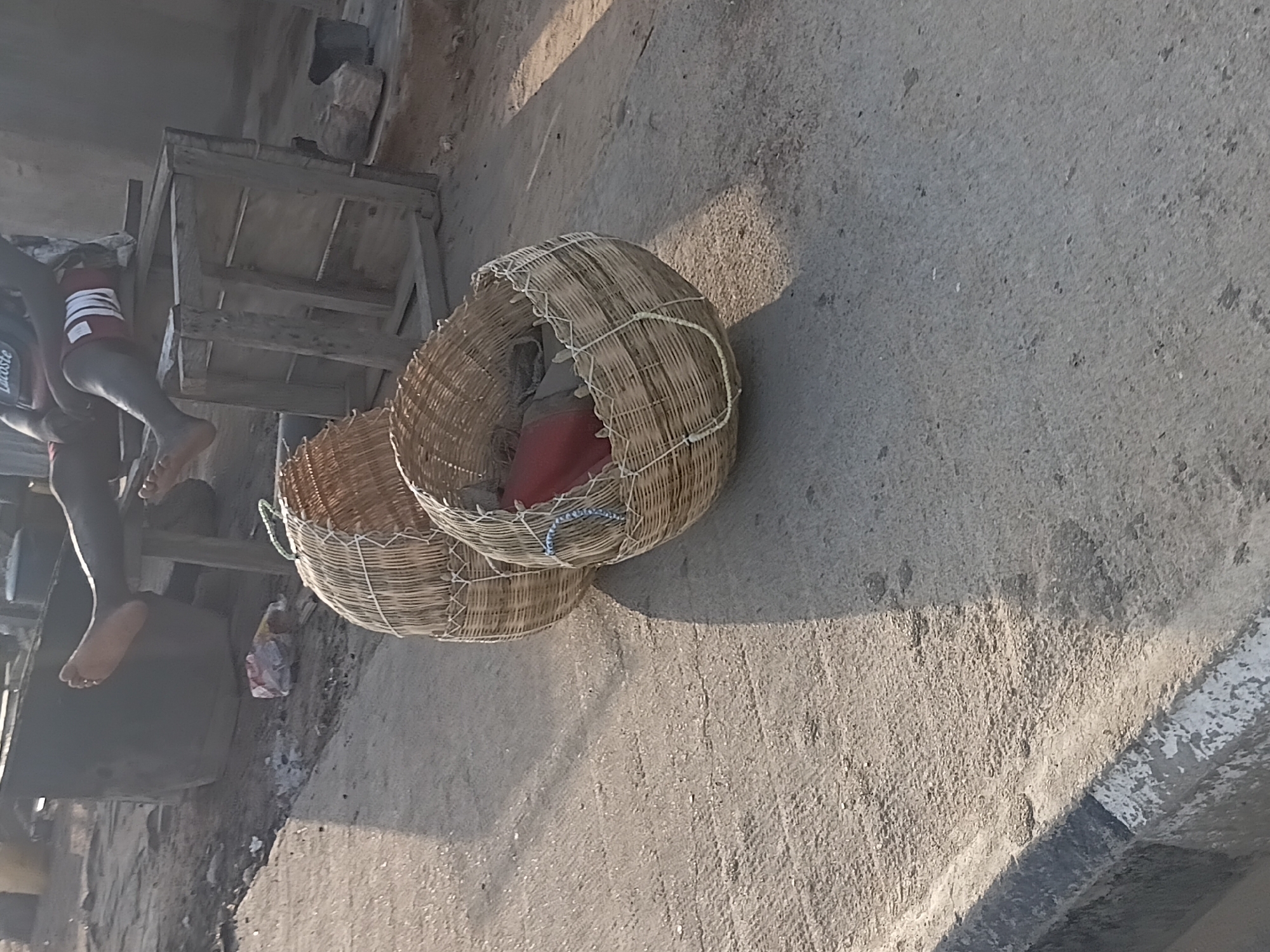 In Jalingo, Nigeria, two woven baskets are situated on a textured concrete surface. One basket, larger and in the foreground, contains a red object and appears to be filled with other items. The second basket, positioned behind and slightly to the left of the first, is also woven and empty. A person's bare feet are visible in the upper portion of the image, resting on a wooden structure. The background is indistinct but suggests an outdoor environment with some large, dark, amorphous objects. The lighting indicates it is daytime, casting shadows on the ground.