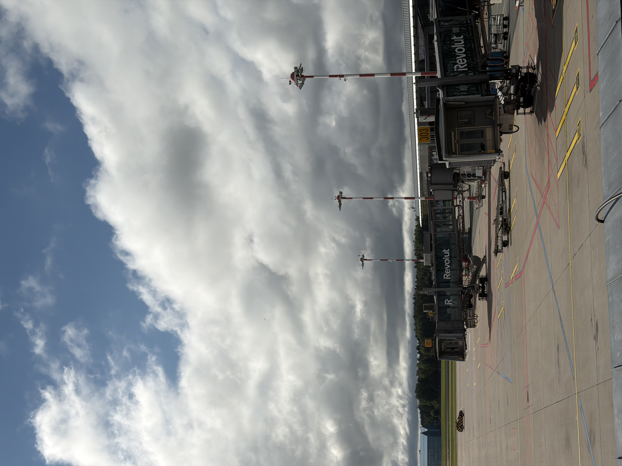The image depicts an airport tarmac in Skulte, Latvia, under a cloudy sky. Several jet bridges extend from a terminal building, with at least one labeled "Revolut." The tarmac is marked with various colored lines and symbols, including yellow crosses. Two striped poles with lights and windsock-like attachments are visible. In the distance, trees and buildings line the horizon. A yellow and black checkered vehicle is parked on the tarmac. The scene is static and appears to be during the daytime.