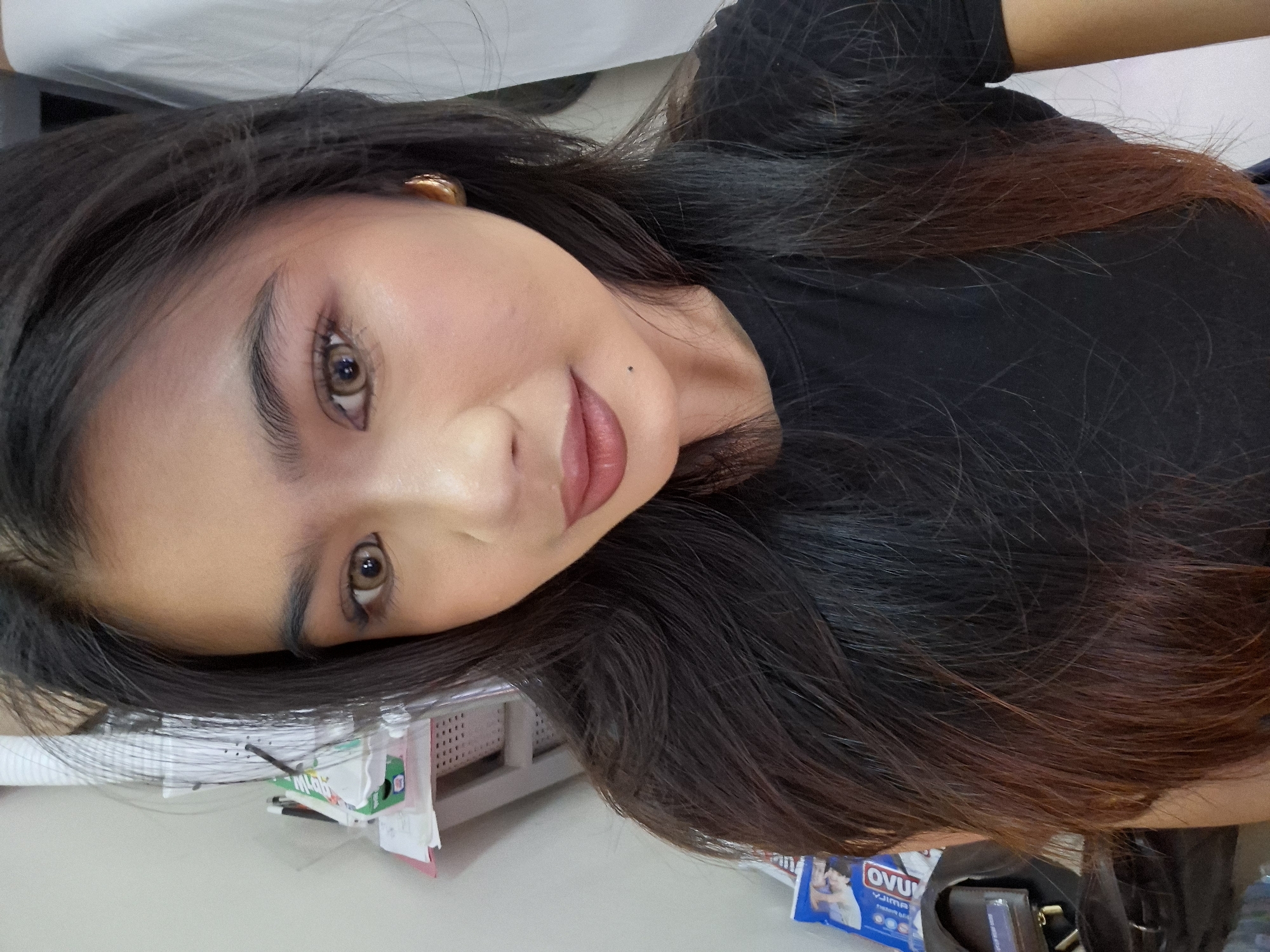A young woman with dark hair and light brown eyes, wearing a black t-shirt, poses for a selfie indoors. She has on makeup, including lipstick and eyeliner, and is wearing gold hoop earrings. Her hair is long and dark brown, with lighter brown highlights towards the ends. The background shows a white wall with some shelving and items on it, suggesting an indoor setting. There is no indication of the city of Kuta, Indonesia, in the image.