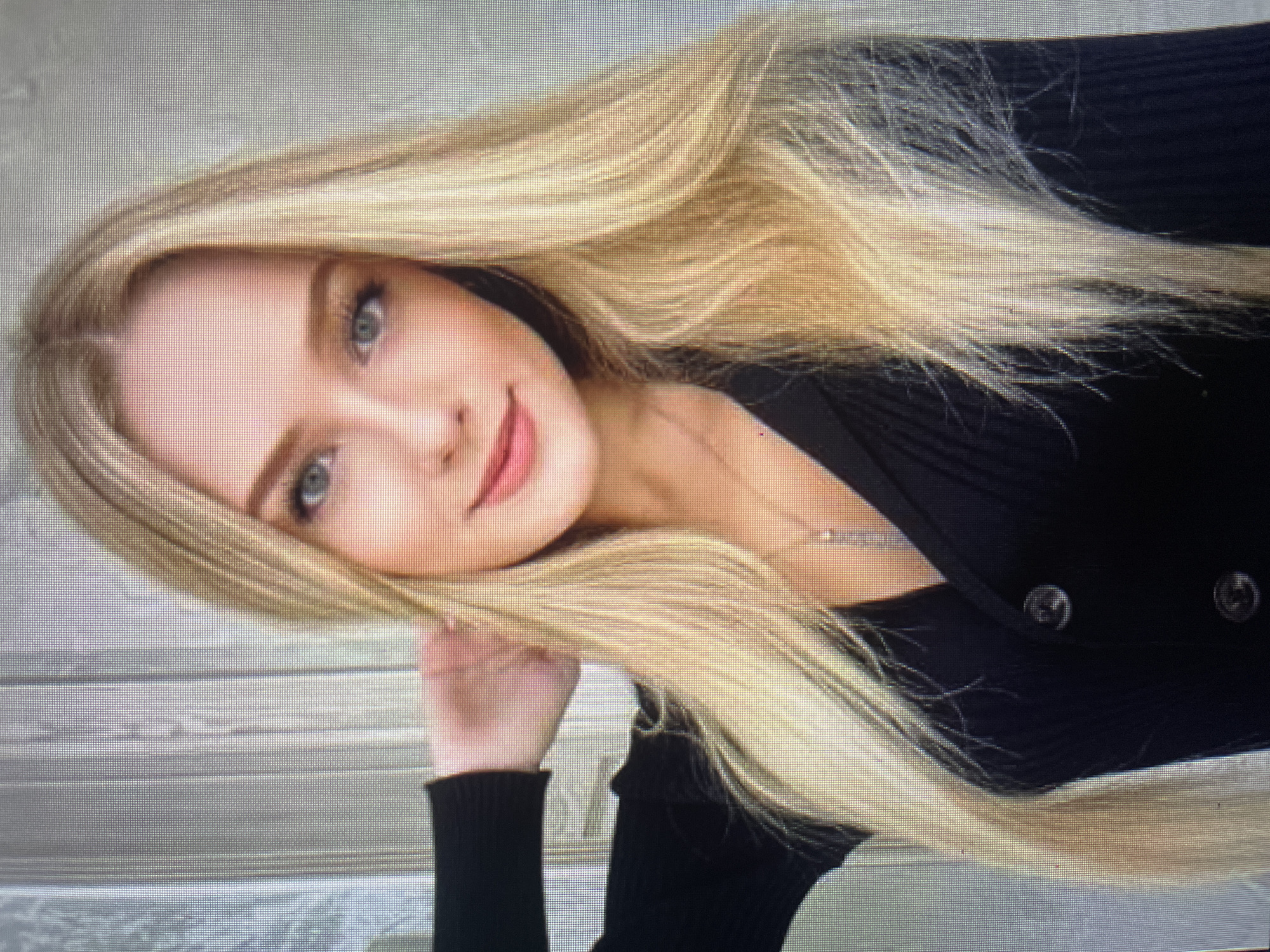 The media file is a photograph of a young woman with long, blonde hair. She is wearing a black top with buttons. Her head is tilted to her right, and her hair is spread across her back and shoulder. She has blue eyes and is smiling. The background appears to be a light-colored, textured surface, possibly wooden slats or blinds. The lighting is bright, suggesting an indoor setting with good illumination. There is no visible text in the image. The overall mood of the photograph is relaxed and pleasant.