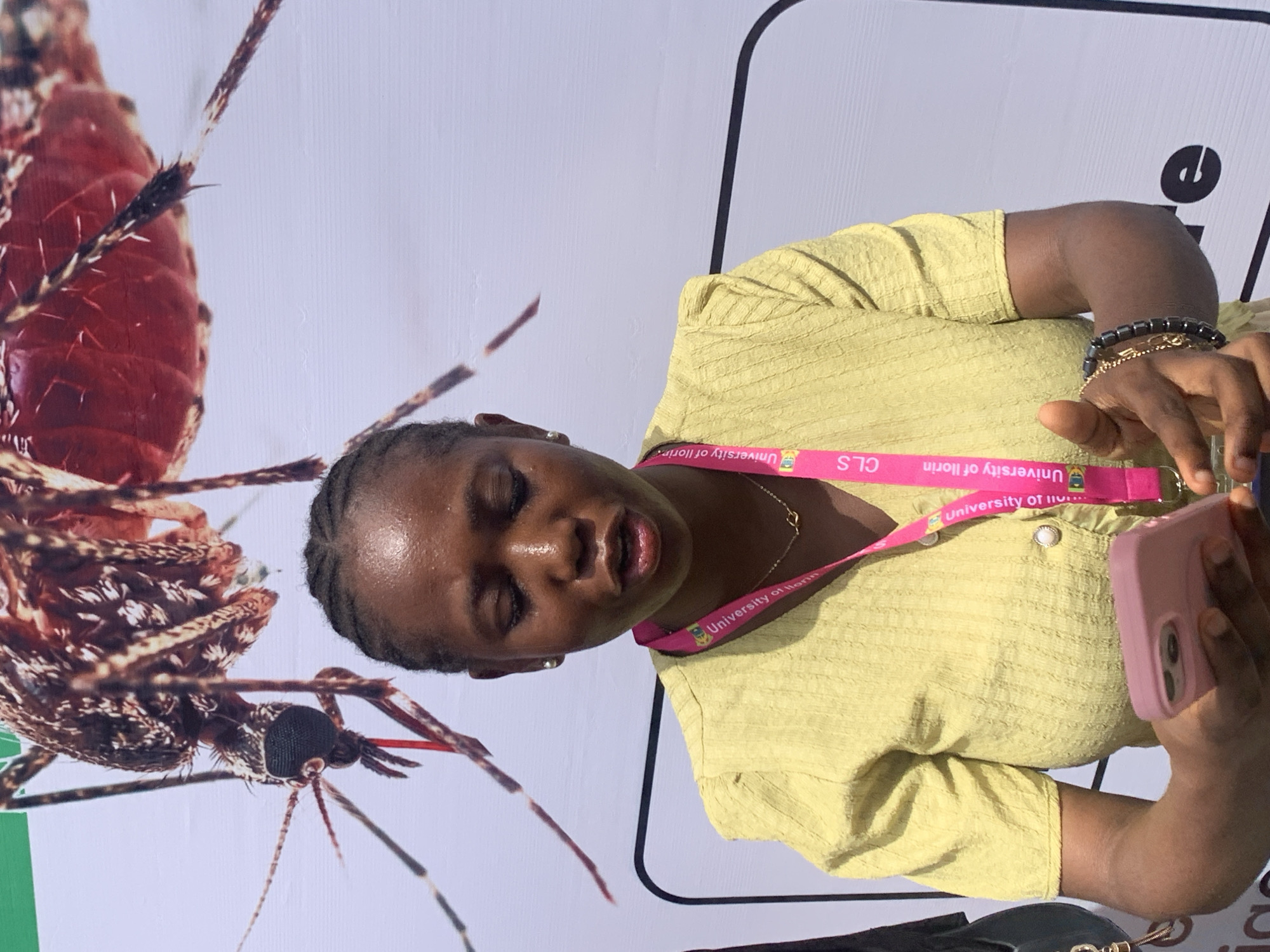 A young Black woman is depicted in a mid-shot, engaged with a pink-cased smartphone held in her hands. She is positioned facing slightly right, looking downwards at her phone with her eyes closed or nearly closed and her mouth slightly open, suggesting a candid moment of concentration or interaction. She wears a light yellow, textured short-sleeved top and a delicate gold chain around her neck. A pink lanyard, clearly visible, bears the repeated text "University of Ilorin" and the letters "CLS." Her right wrist is adorned with several bracelets, including one made of dark, rectangular beads.
Behind her is a large white banner or poster featuring a highly magnified, detailed image of a reddish-brown mosquito. The insect's segmented body, intricate patterns, fine hairs, legs, large compound eyes, and proboscis are distinctly visible. A narrow green stripe is partially seen at the bottom left of the banner, and a bold black curved line is present towards the top right, likely part of a graphic design.
The setting appears to be an event or educational environment, possibly related to public health or entomology, given the prominent mosquito imagery and the affiliation with the University of Ilorin, located in Ilorin, Nigeria. The bright, even lighting suggests a daytime setting, either indoors or in a sheltered outdoor space.