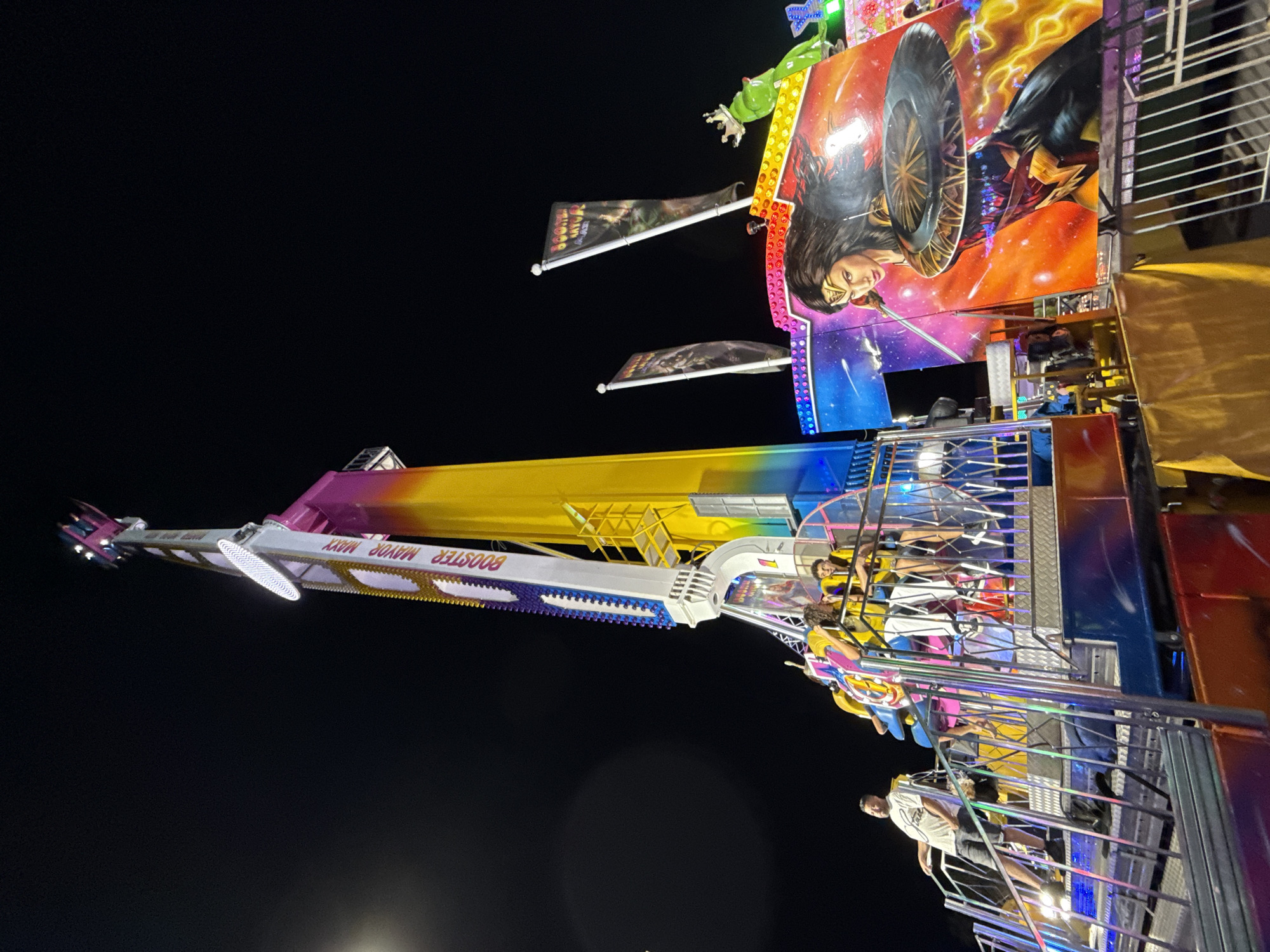 An extreme ride, possibly a drop tower or swing ride, is illuminated at night in what appears to be a fairground or amusement park area in Blanes, Spain. The ride's tall, slender tower is painted with a rainbow gradient, transitioning from yellow at the bottom to pink at the top, with white and purple accents. Several gondolas are attached to the tower, and some are occupied by people wearing safety harnesses.
In the foreground, a person in a white t-shirt and grey shorts stands on a platform, looking to the left. Behind them, on the ride's platform, other individuals, some wearing yellow, are seated in the gondolas. The ride structure features a prominent graphic of Wonder Woman and possibly Batman against a cosmic background, adorned with colorful LED lights. Two flags with an image of a knight on horseback are also visible, one partially obscuring the other. A neon green frog decoration with a crown sits atop the ride's facade. The scene is set against a dark night sky, with the moon visible as a faint, luminous orb in the upper left quadrant. The overall impression is one of an active amusement park at night, with the ride as the central focus.