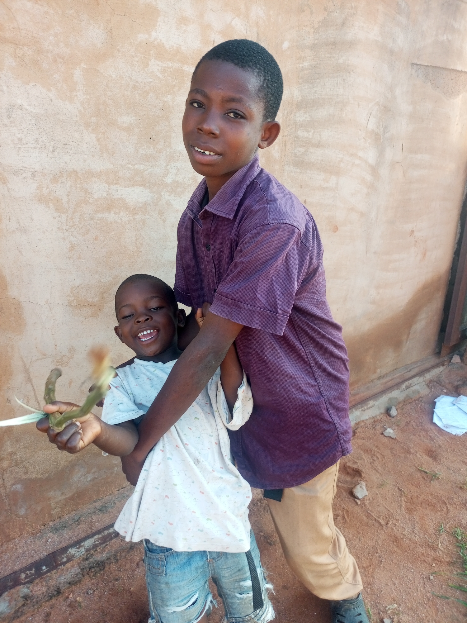 Two young Nigerian boys are pictured outdoors in Tempure, Nigeria, against a textured, light-colored wall. The older boy, wearing a purple collared shirt and tan pants, stands behind and to the right of the younger boy, embracing him. The younger boy, in a light blue shirt with a pattern and ripped denim shorts, faces forward and smiles broadly, holding a green plant stem in his left hand. The ground appears to be a mix of dirt and sand with some scattered stones and vegetation. A folded white cloth is visible to the far right of the image.