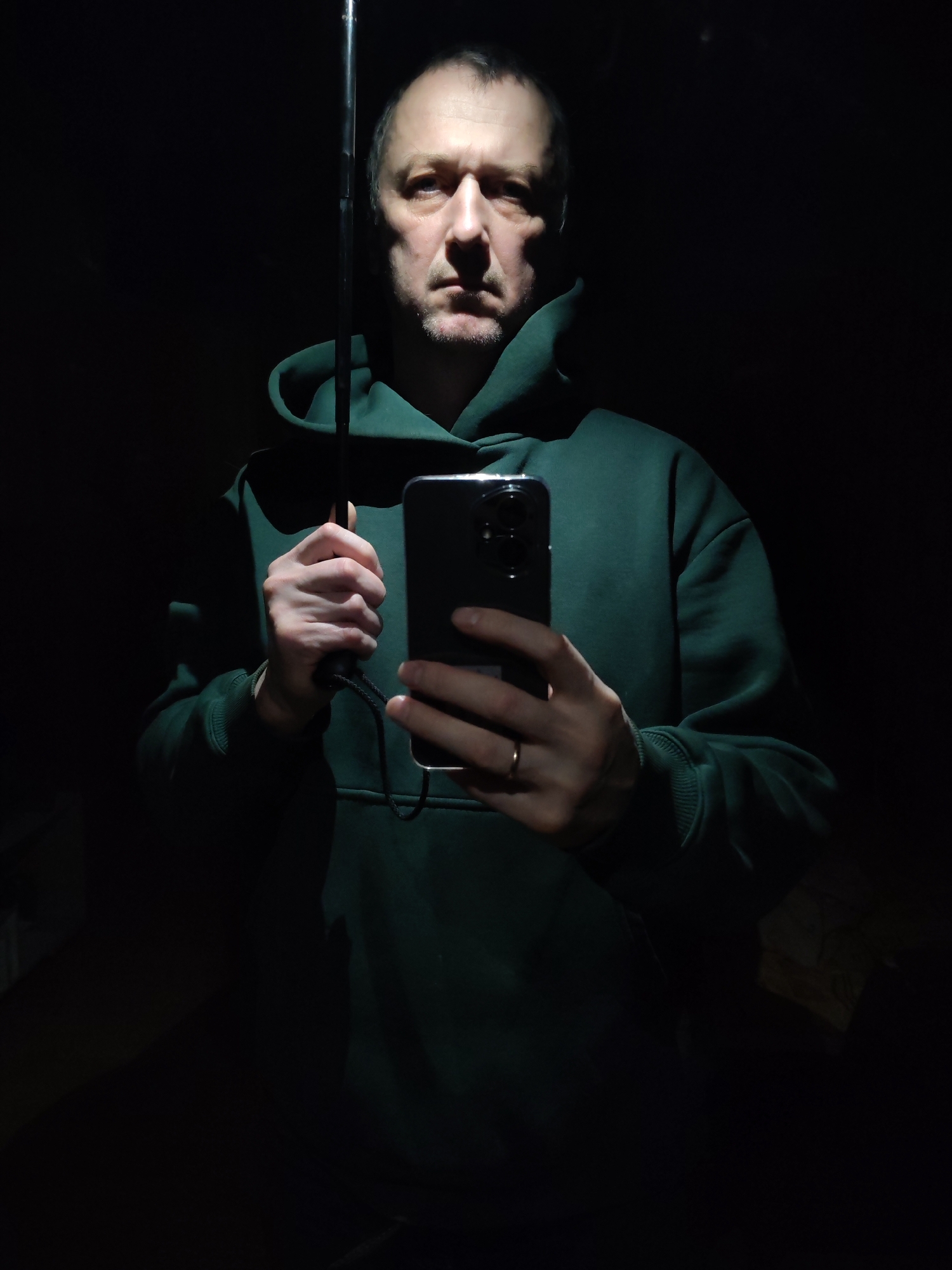 A medium shot shows a man in a dark green hooded sweatshirt holding a black smartphone and what appears to be the handle of an umbrella. The man has short, dark hair, and his face is lit from below, casting harsh shadows. He has a stern expression. He is wearing a ring on his left hand. The background is dark and indistinct. There is no indication in the image that pinpoints the location to the city of Chaikovsky, Russia.