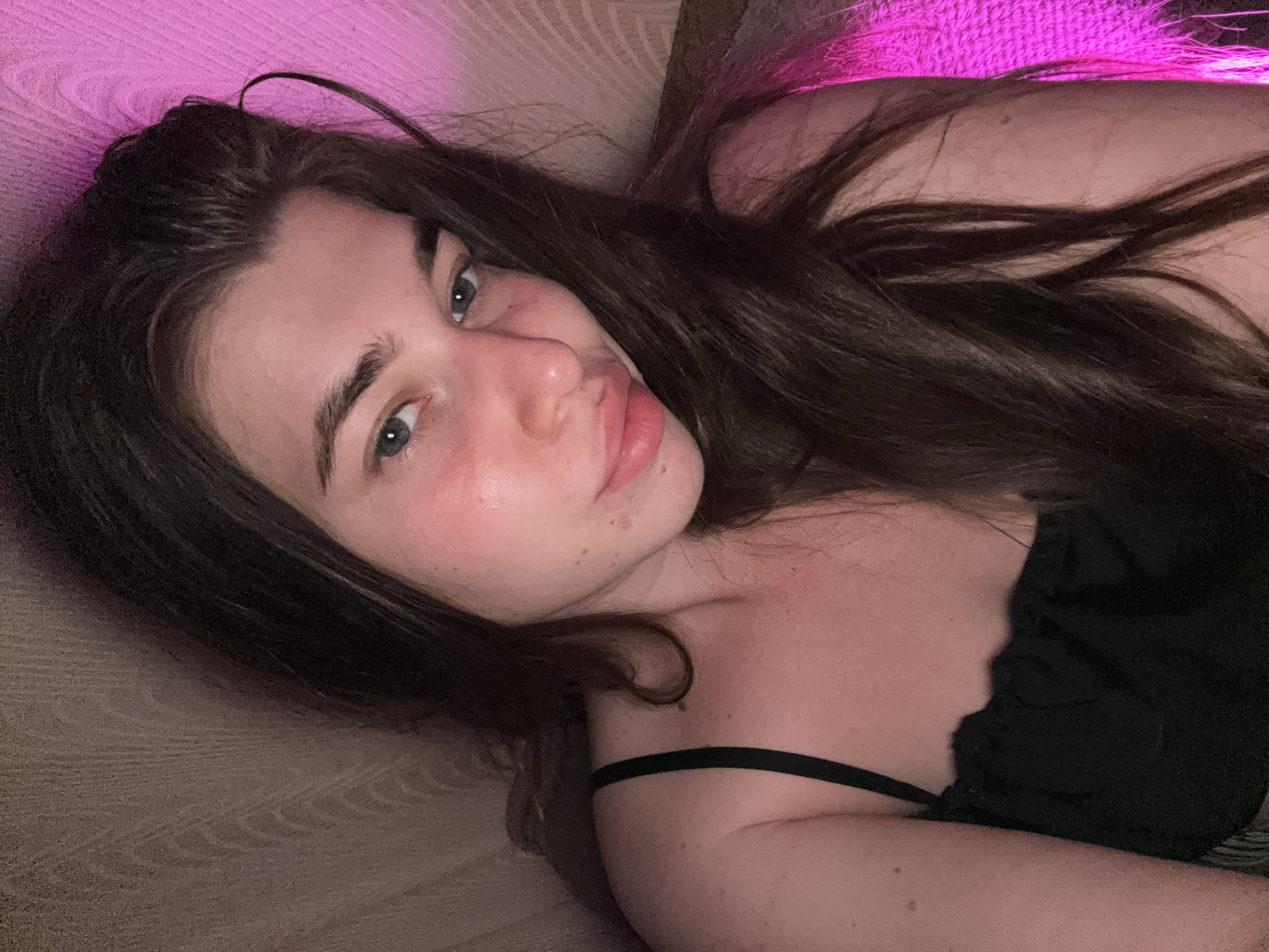 A young woman with long, dark brown hair is lying down, with her head resting on a textured surface. She is wearing a black camisole. Her face is turned towards the camera, with her left eye visible and her right eye partially obscured by her hair. There is pink lighting in the background.