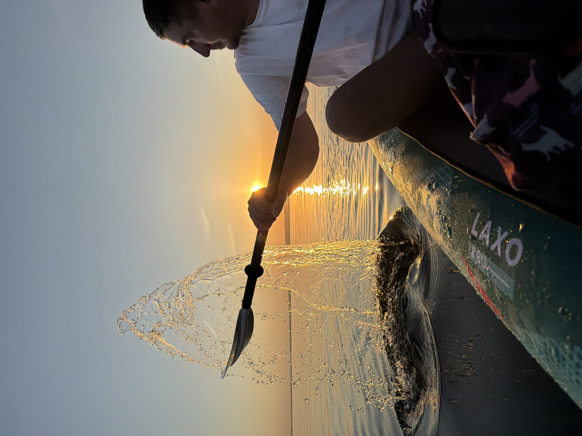 The photograph shows a person paddling a stand-up paddleboard (SUP) at sunset. The sun is low on the horizon, creating a bright glare and golden light on the water. The paddler, seen from the side, wears a white shirt and holds a paddle with their hand obscuring the sun. Water splashes near the paddleboard. The SUP is green with the word "LAXO" visible on the side. The location is unknown and cannot be determined from the image.