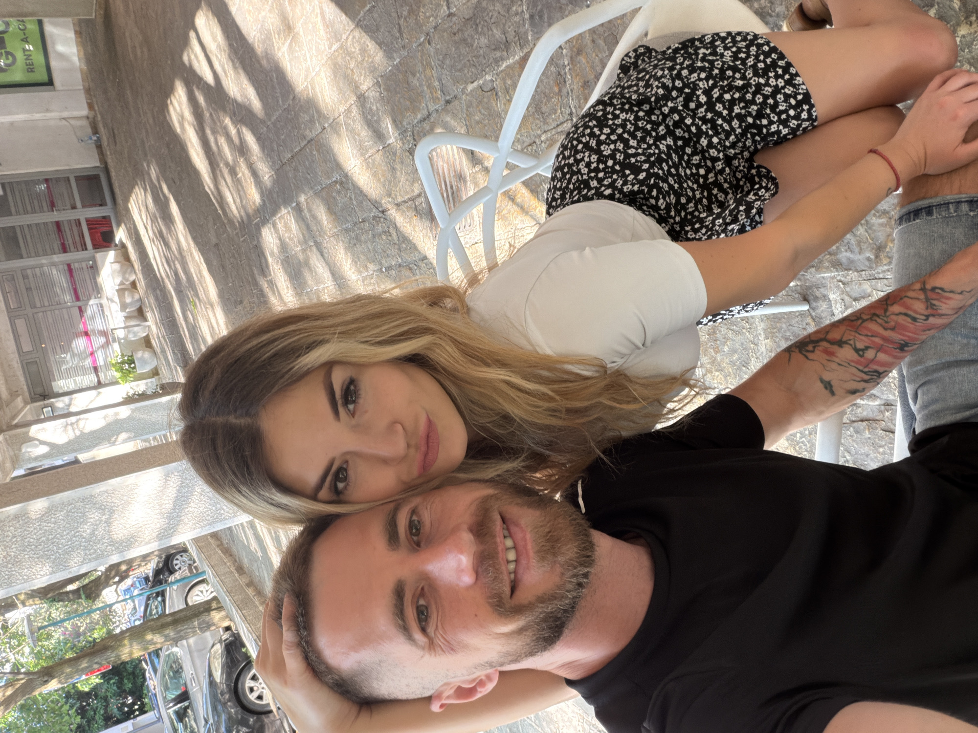 A young Caucasian couple is taking a selfie outdoors, likely in Budva, Montenegro, given the presence of a sign that reads "RENT-A-CAR." The man, with short brown hair and a beard, is in the foreground on the left, smiling and looking at the camera. He is wearing a black t-shirt and has a tattoo on his left arm. The woman, with long blonde hair, is leaning on his shoulder on the right, also looking at the camera and pouting her lips. She is wearing a white t-shirt and a black and white floral skirt. They are seated on white chairs at an outdoor seating area, with a stone-paved ground and shadows from trees and structures falling on it. In the background, there are buildings with glass doors and windows, some potted plants, and parked cars. The lighting suggests it is daytime, possibly late afternoon, with sunlight casting distinct shadows.