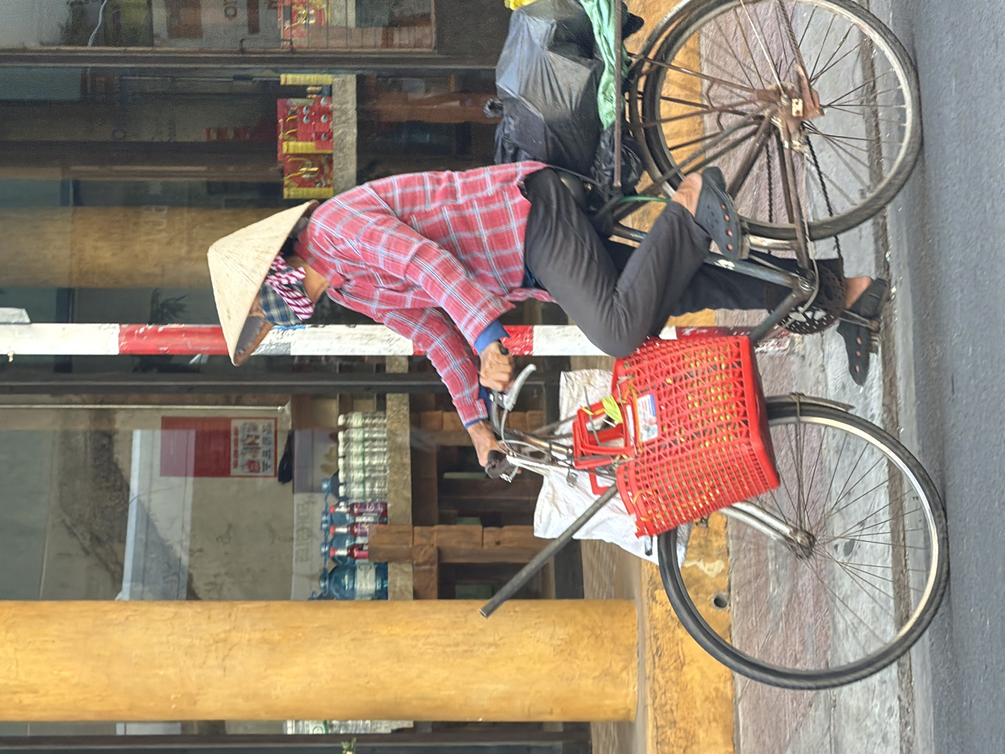 A person wearing a conical hat and a plaid shirt rides a bicycle laden with goods. The bicycle has a red basket in the front and a black trash bag and green cloth in the back. The person is wearing dark pants and black clogs. The scene appears to be on a street in Nha Trang, Vietnam, with buildings and shops in the background. A yellow pillar is on the left side of the image. The sun is shining, casting shadows.