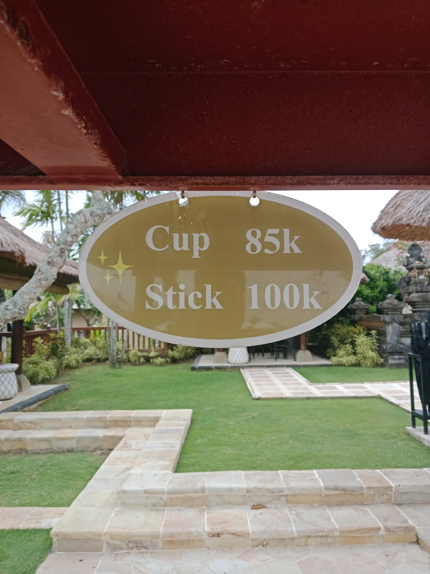 This image shows an oval sign with a golden-yellow background and a white border. The sign is hanging from a reddish-brown structure. There are three yellow starbursts on the left side of the sign. The text on the sign is in white capital letters. The top line reads "Cup 85k" and the bottom line reads "Stick 100k".
The sign is placed outdoors in a lush green setting. In the background, there are palm trees, thatched-roof structures resembling huts or gazebos, and what appears to be traditional Balinese stonework, including a statue or shrine. A paved pathway winds through the grass. The overall impression is of a tropical resort or garden area.
The lighting suggests it is daytime, and the weather appears to be clear. The image is taken from a low angle, looking up towards the sign. There are no people visible in the image. The scene conveys a sense of relaxation and vacation. The location is identified as Kuta Selatan, Indonesia, which is consistent with the visual cues of tropical foliage and Balinese architecture.