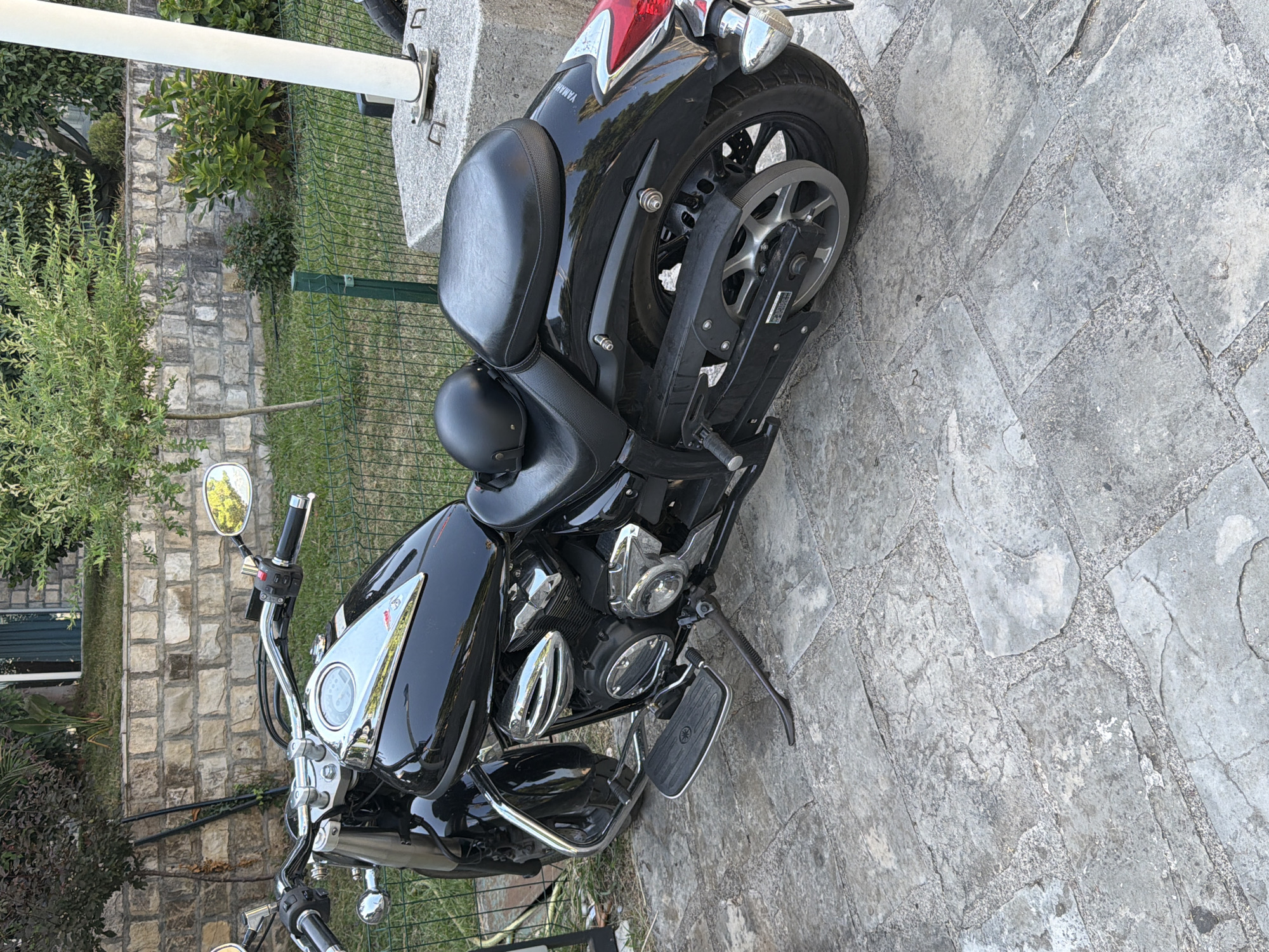 A black Yamaha motorcycle is parked on a stone patio in Budva, Montenegro. The motorcycle is a cruiser-style bike with chrome accents. It has a black leather seat, a black helmet resting on the seat, and a black exhaust pipe. The motorcycle is in good condition and appears to be well-maintained. The background shows a stone wall, greenery, and a fence. The lighting suggests it is daytime.