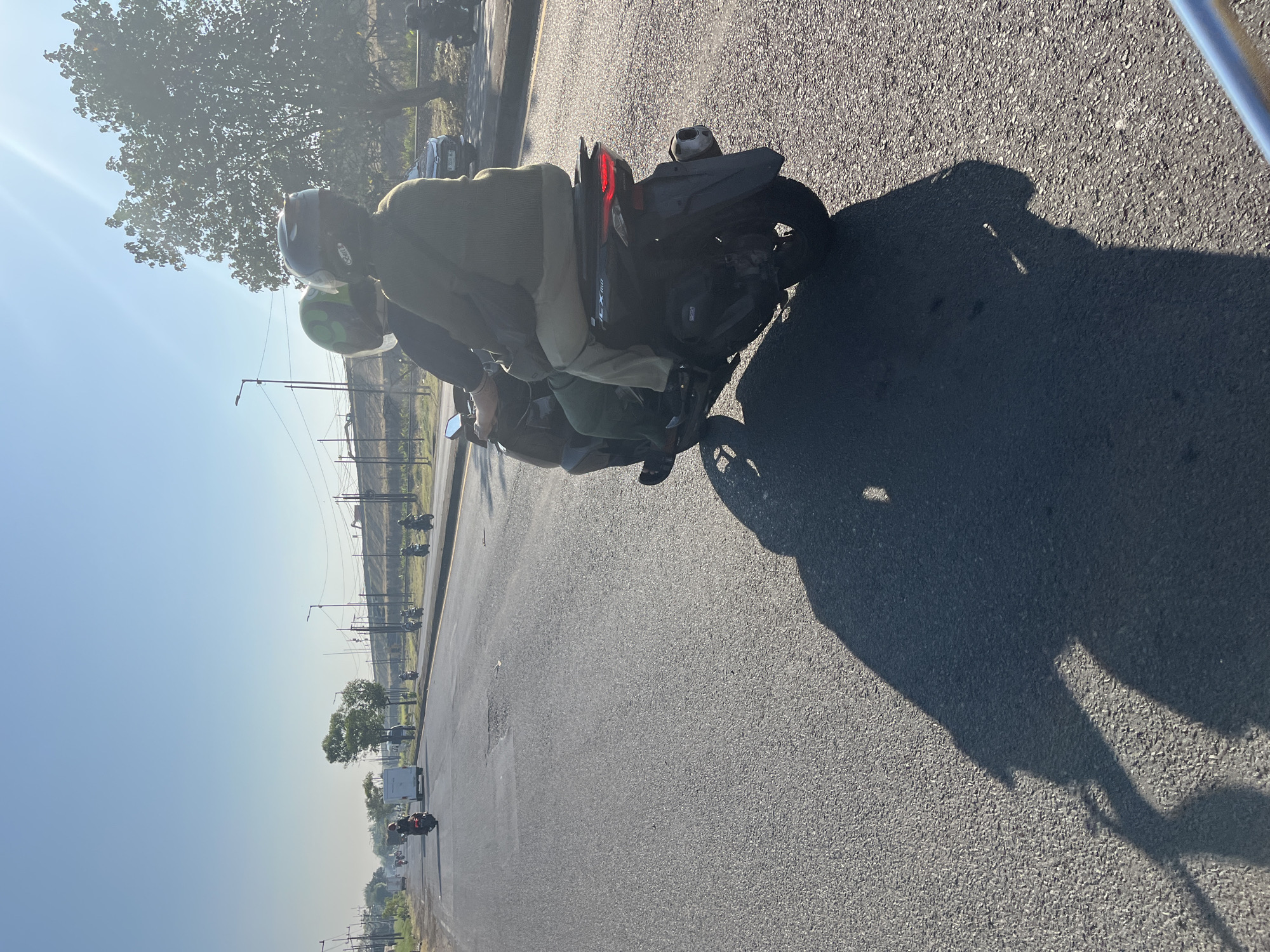 In Ketapang, Indonesia, on a sunlit day, two individuals ride a dark-colored scooter down a paved road. The person in the rear, wearing a green jacket and light-colored pants, is driving. The passenger in front wears a green helmet with a black pattern and a light-colored jacket. Both individuals have helmets on. The scooter's shadow stretches across the road, indicating the strong sunlight. In the background, other vehicles, including motorcycles and a white van, travel on the same road. Trees and utility poles line the side of the road, and a bright, clear sky suggests fair weather. The road surface appears worn in places, with a visible pothole.