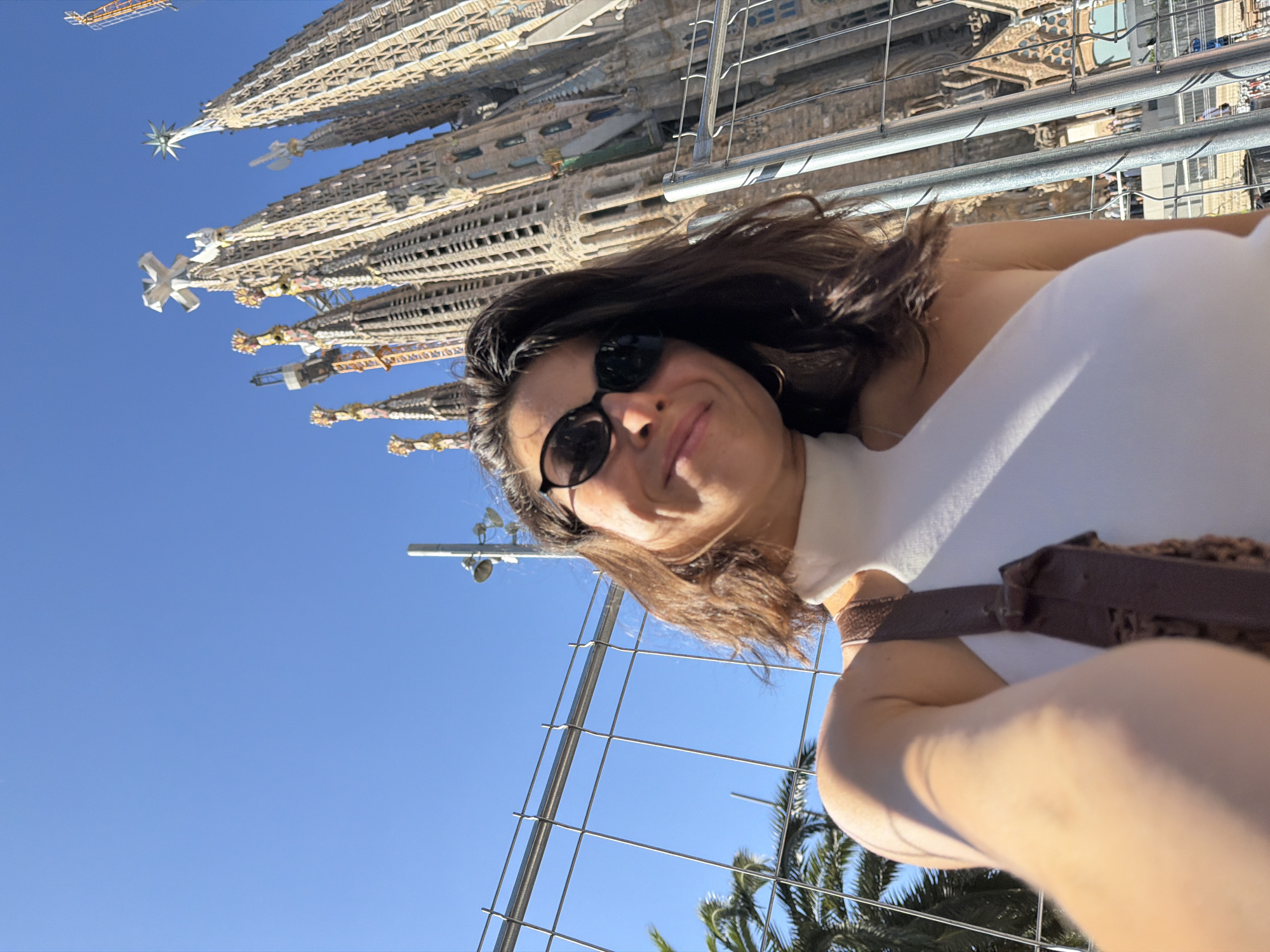 A low-angle, wide-shot selfie shows a woman in Barcelona, Spain, in front of the Sagrada Familia. The woman, facing the camera, wears a white one-shoulder top and black sunglasses. She has shoulder-length dark brown hair and is smiling. A brown purse strap is visible across her shoulder. Behind her, the iconic spires of the Sagrada Familia dominate the background, appearing under construction with scaffolding present. The sky is a clear, vibrant blue. A metal fence runs across the foreground, partially obscuring the view of palm trees to the left of the frame.