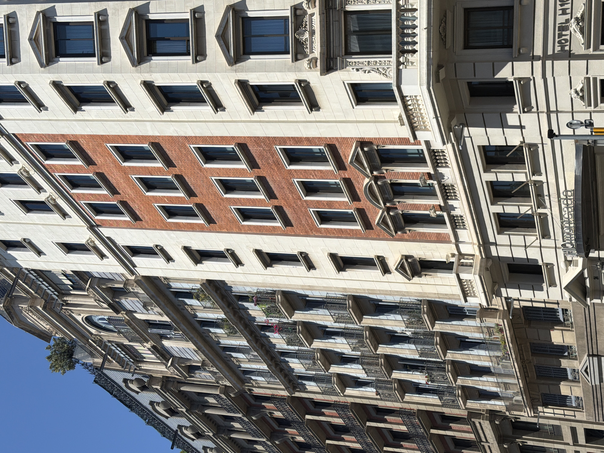 The image shows a tall building with a mix of architectural styles. The facade is predominantly white with rows of windows, some of which have decorative elements above. There's a section with red brick and similar windows, creating a contrast. A portion of the building has balconies with railings.
The building is likely in Barcelona, Spain. The facade contains the words "Hotel H10" and "Moncada."
The environment is sunny. There are no visible people or actions.