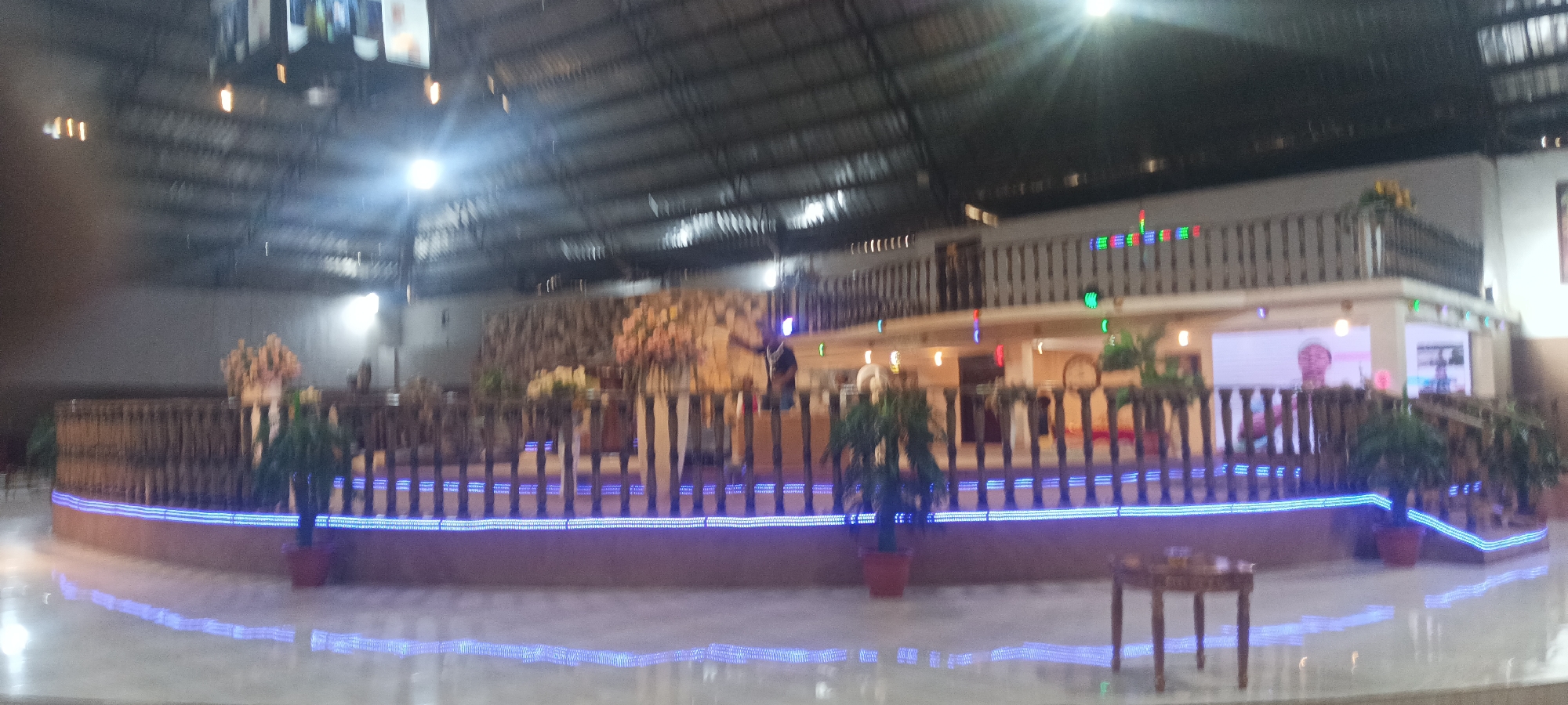 The image depicts the interior of a building, possibly a hall, located on Rukuba Road, Nigeria. The hall has a large stage area with a wooden railing and blue LED lights running along the bottom. Potted plants are positioned along the stage's edge. Behind the stage, there appears to be a decorative wall and a second-story balcony with railing.
Illumination comes from overhead lighting, with some light reflecting off the floor. There is a table with three legs in the foreground. Several people are standing behind the wooden railing on the stage. Behind those people are LED display screens showing a face and a picture of two people.